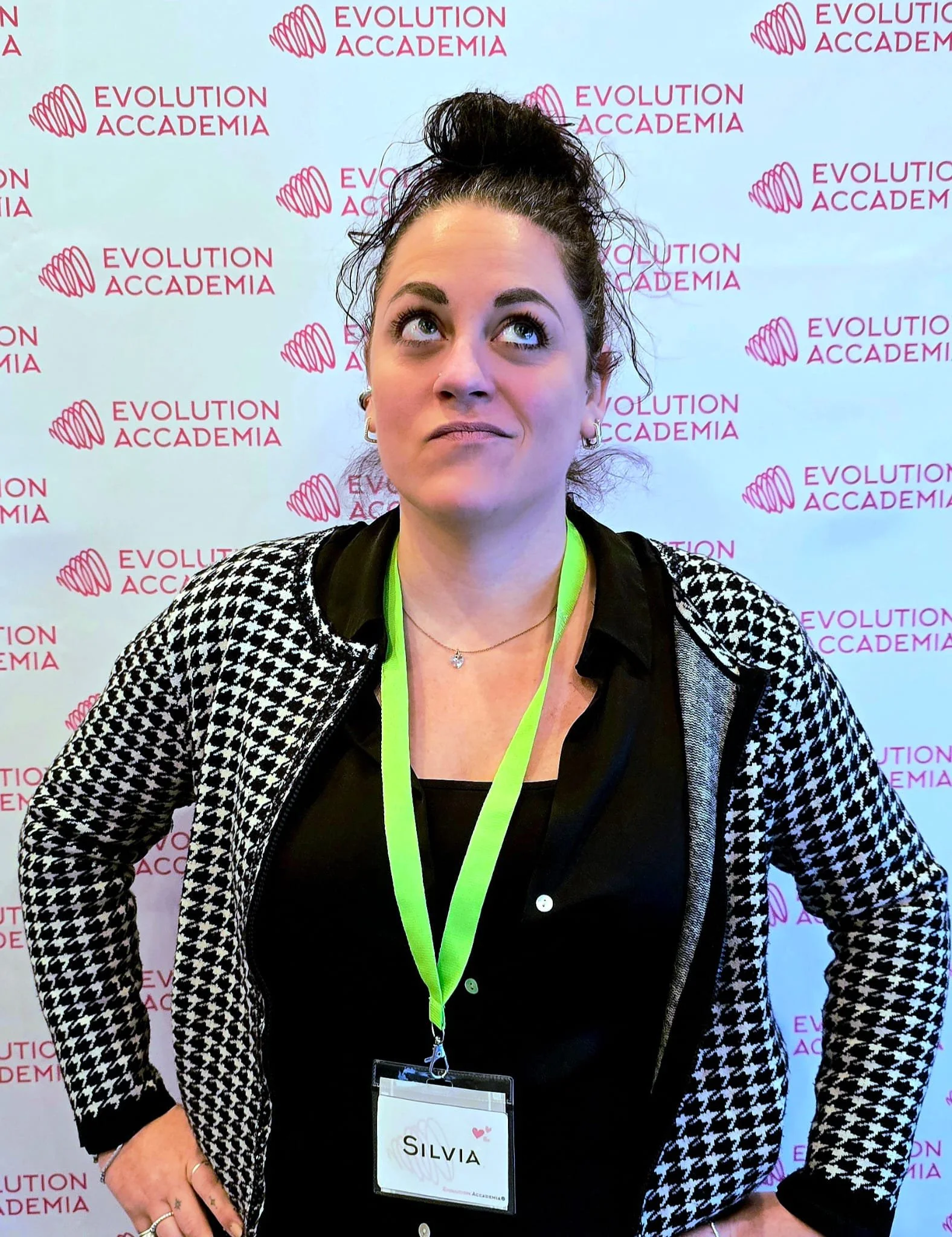 Una donna con i capelli castani raccolti in uno chignon, indossa una giacca a rete con motivo houndstooth e una fascia verde con una targhetta con il nome Silva, davanti a uno sfondo con il logo di Evolution Accademia.