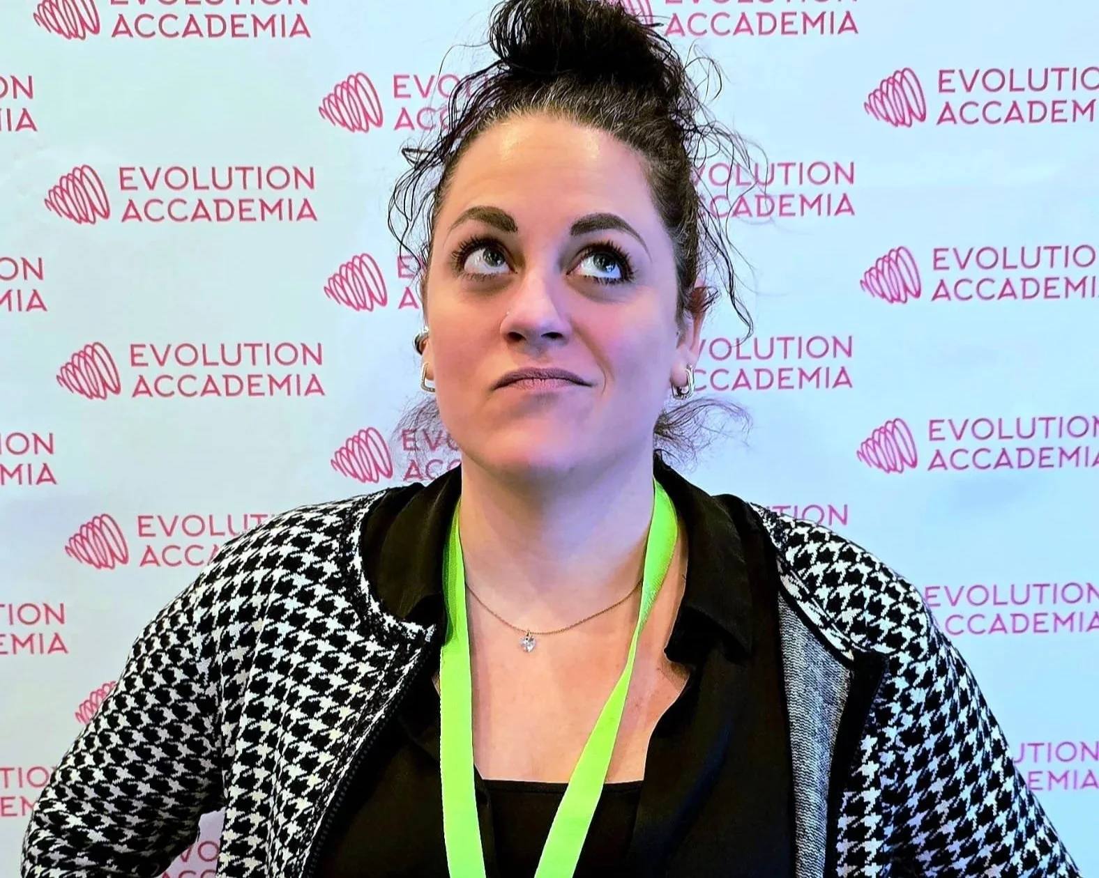 Una donna con i capelli raccolti in una treccia alta e occhi azzurri, indossa una maglietta nera, una giacca con motivo a scacchi e una collana con ciondolo. Sta davanti a uno sfondo bianco con il logo e il testo 'Evolution Accademia' in rosa.