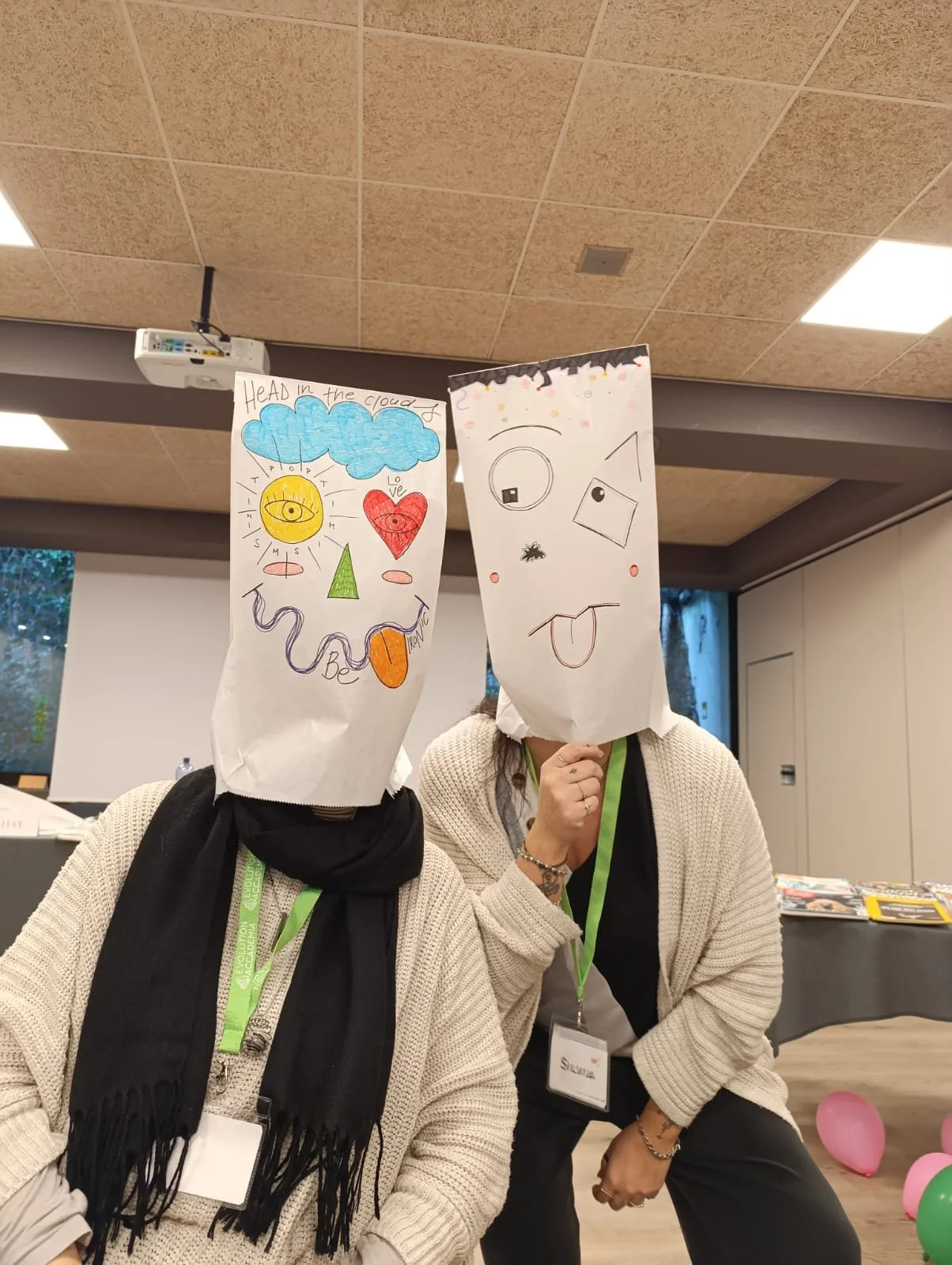 Due persone con teste di carta decorate con disegni, una con un volto con un sole e una nuvola, l'altra con un volto con occhi disegnati, lingua fuori e piccoli disegni intorno.