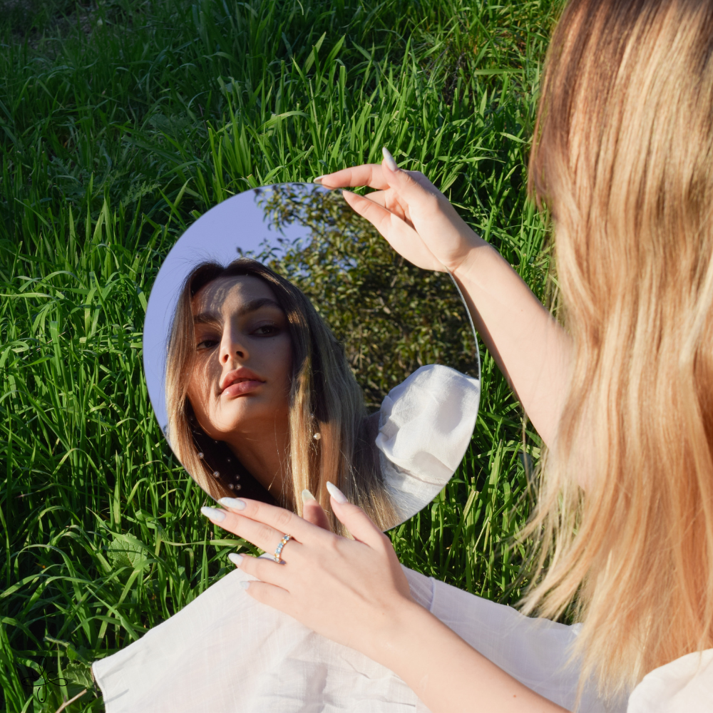 Una donna con i capelli biondi tiene uno specchio rotondo riflettente tra le mani, guardando il proprio volto riflesso, mentre si trova in un campo di erba verde.