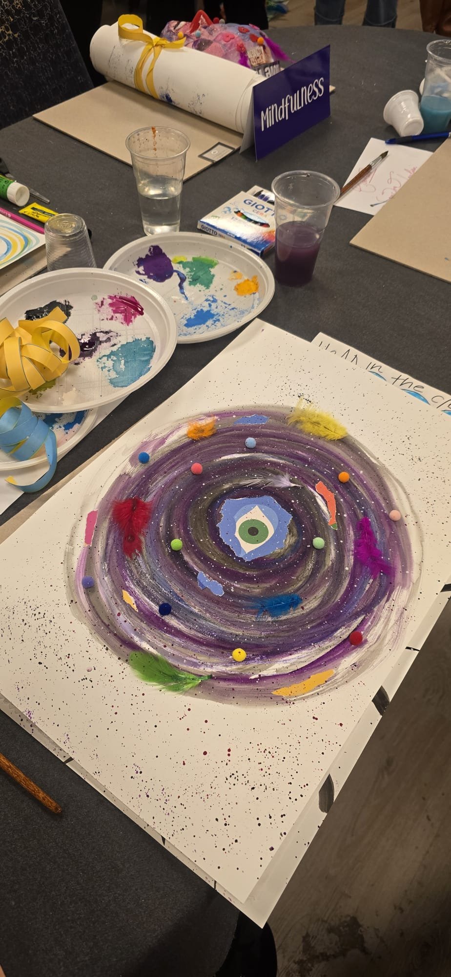 Un dipinto astratto con un motivo centrale che ricorda un occhio, circondato da colori vivaci e piccoli oggetti colorati, su un foglio bianco. Sul tavolo ci sono piatti con colori, pennelli e altri materiali artistici.