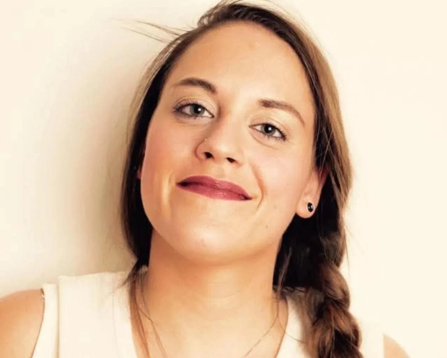 Ragazza con capelli castani e una treccia, sorridente, indossa una maglietta bianca e ha orecchini neri, sfondo chiaro.