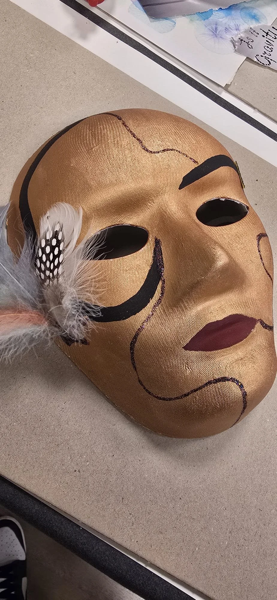 Maschera da teatro dorata con dettagli neri e un fiore di piume bianco e nero sulla sinistra.