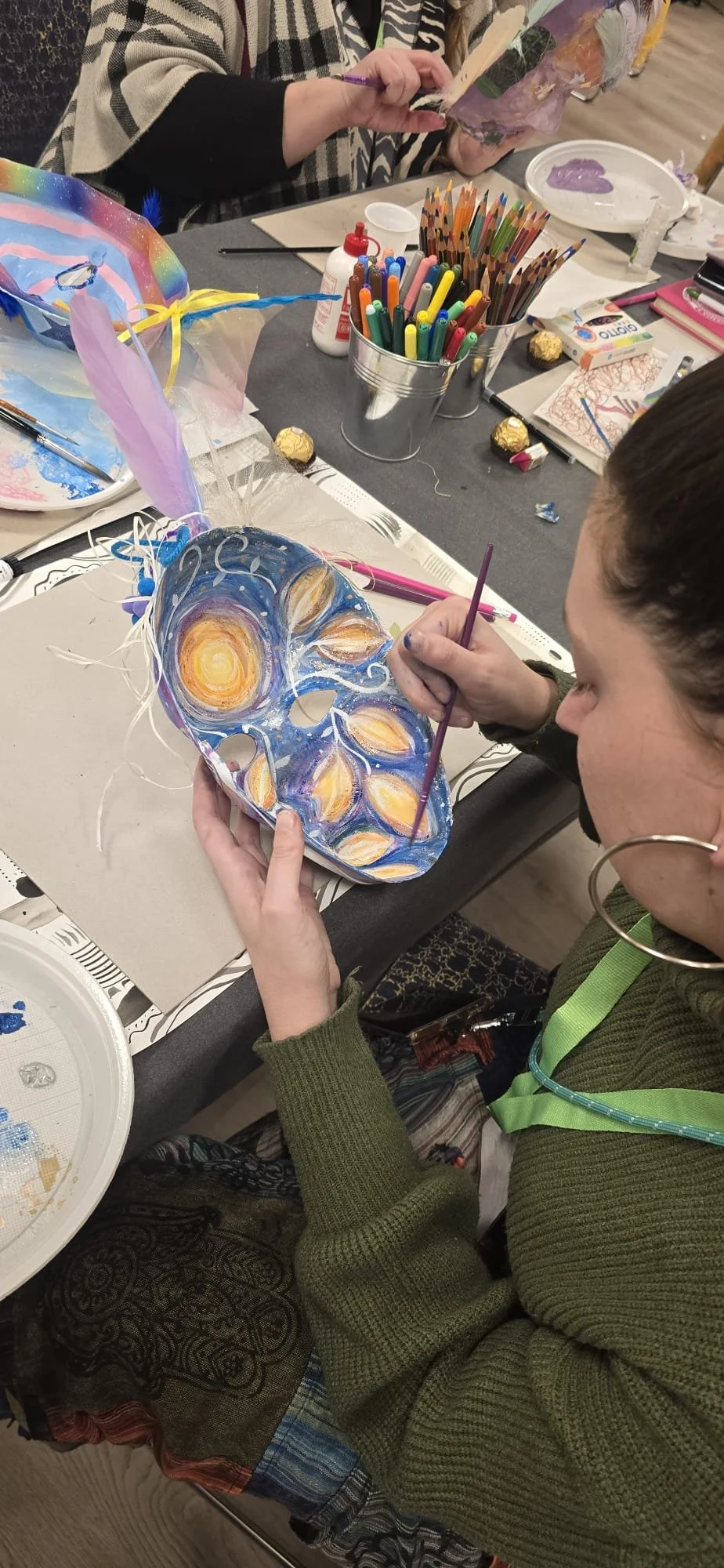 Una donna che dipinge con stili ispirati a Van Gogh su una piastra decorativa, seduta a un tavolo con vari strumenti e materiali artistici.