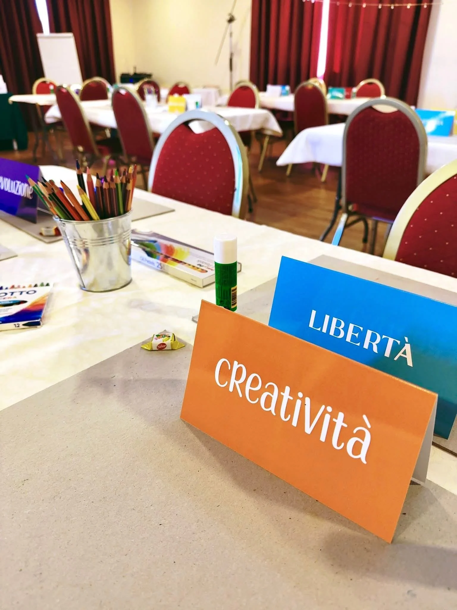 Sala riunioni o aula con tavoli e sedie rosse, materiale per attività creative come colori, collage e colla, cartelloni con parole italiane come 'Libertà' e 'Creatività' sulla scrivania.