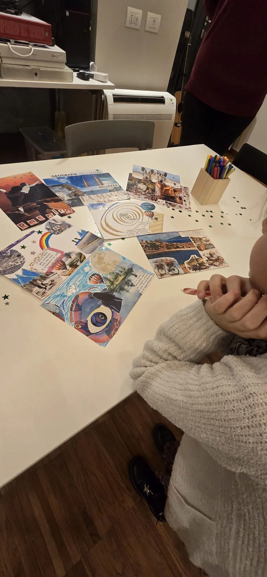 Tabella rotonda con disegni e fotografie colorate su un tavolo, tra cui paesaggi, animali e scene di viaggi, con pennarelli in un porta oggetti di legno e una persona con le braccia incrociate che indossa un maglione beige.
