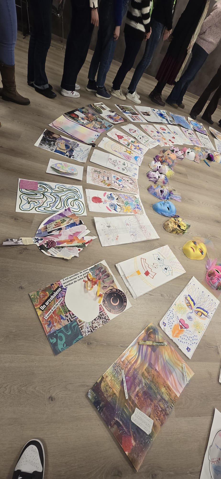 Disposizione di vari disegni, collage e maschere artistiche su un pavimento di legno, con persone in piedi sullo sfondo.