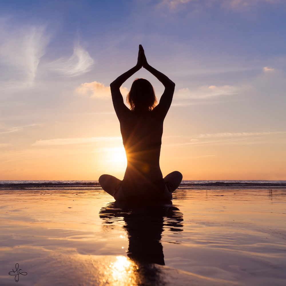 Una persona seduta in posizione di meditazione sulla spiaggia durante il tramonto, con le mani unite sopra la testa e il sole che tramonta sul mare.