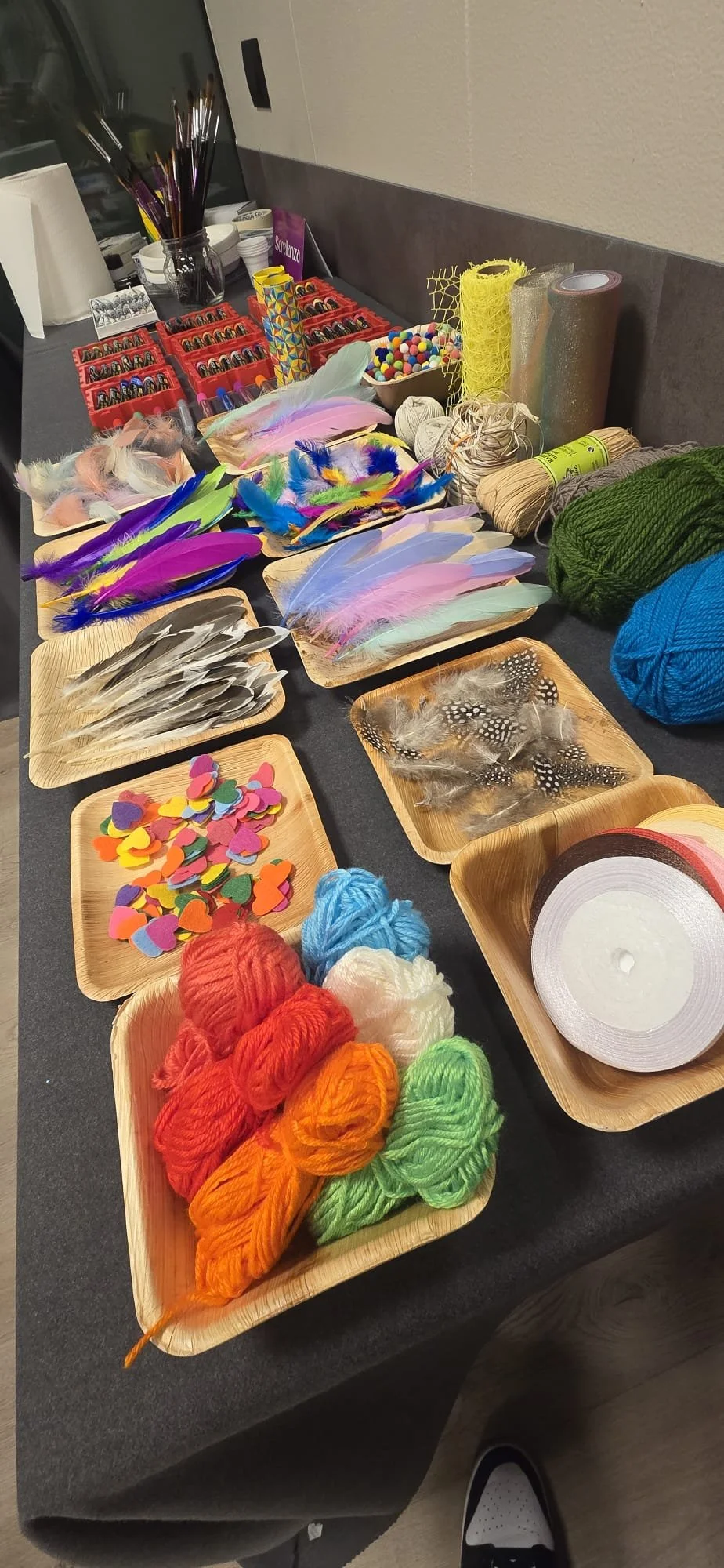 Kit di strumenti e materiali per bricolage o decorazioni, inclusi penne colorate, piume, nastri, cordicelle, bottoni, pezzi di feltro colorati, fiocchi adesivi, bobine di spago, rotoli di nastro adesivo, e filo di lana di vari colori