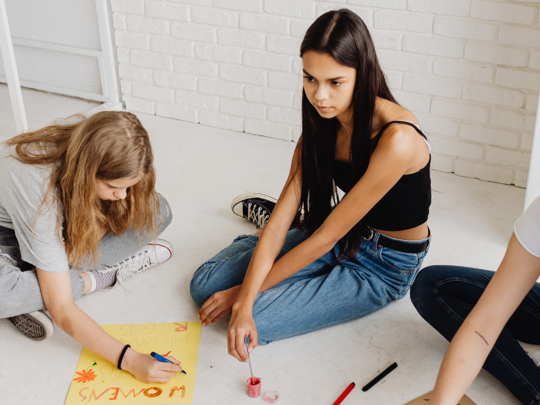 Tre ragazze siedono sul pavimento mentre decorano un cartellone giallo con pittura e penne. Una di loro sta scrivendo e le altre due la osservano.