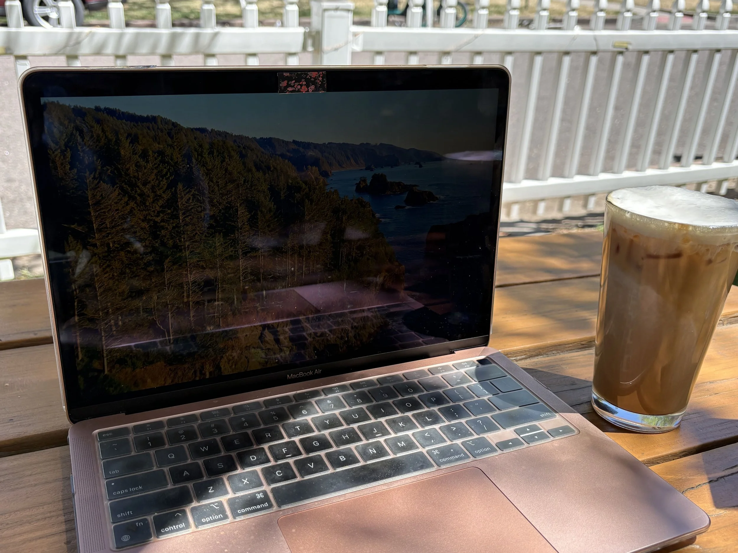 A laptop on a wooden table near a window with a cup of iced coffee beside it, showing a scenic landscape on the screen with trees and water.