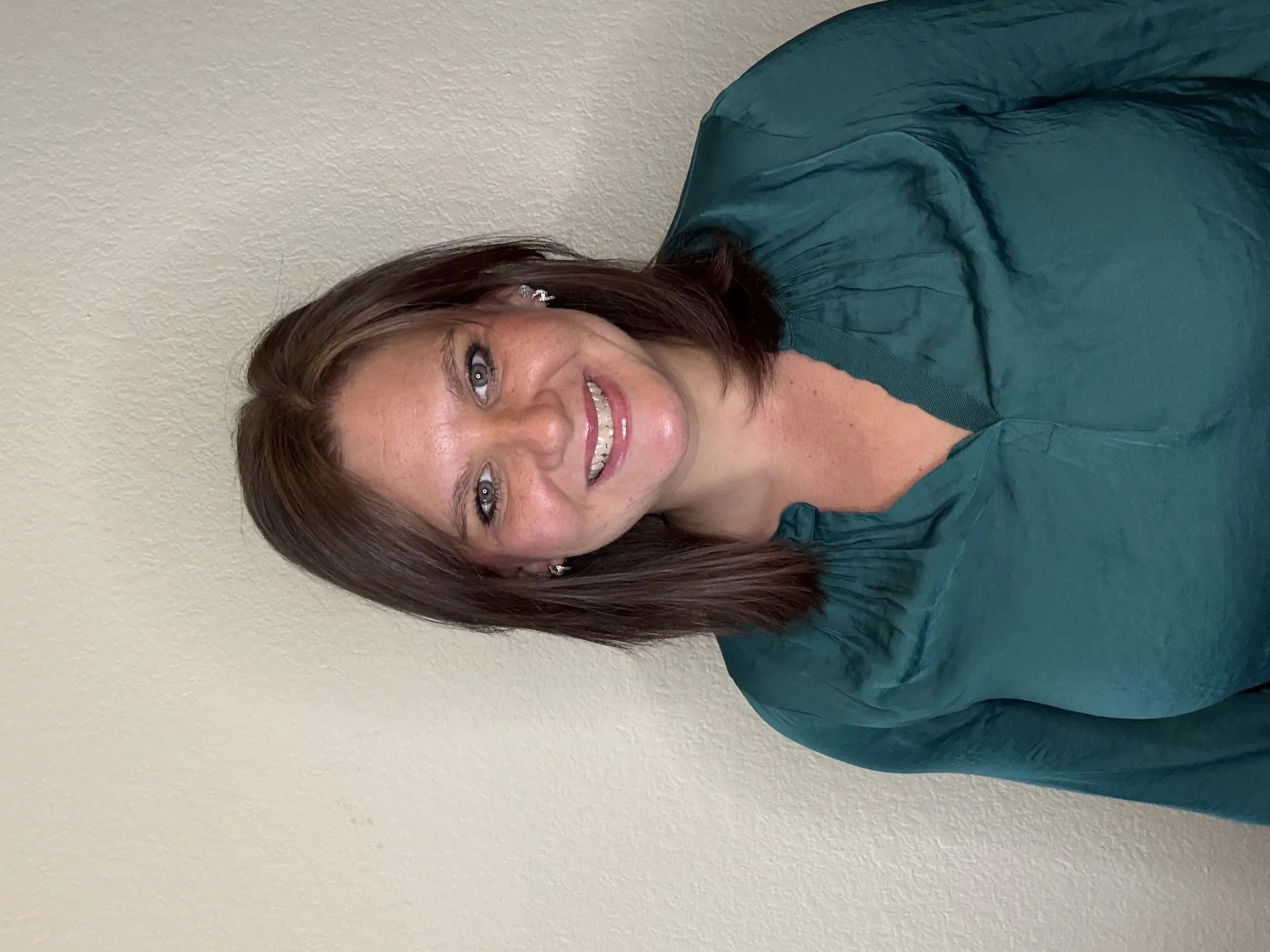 A woman with shoulder-length brown hair, wearing a teal shirt, smiling and standing in front of a plain off-white wall.