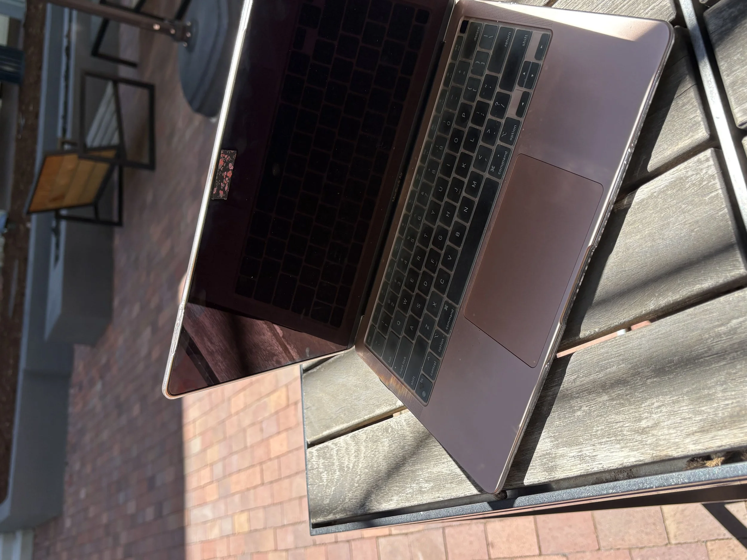 A silver laptop open on a wooden table outdoors, with black keys and a trackpad, next to a glass of water and a small jar.