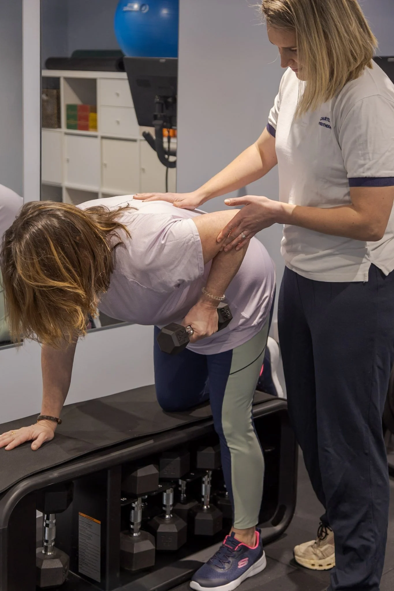 A woman is doing a plank while holding dumbbells, with another woman assisting her and a trainer nearby, in a gym setting.