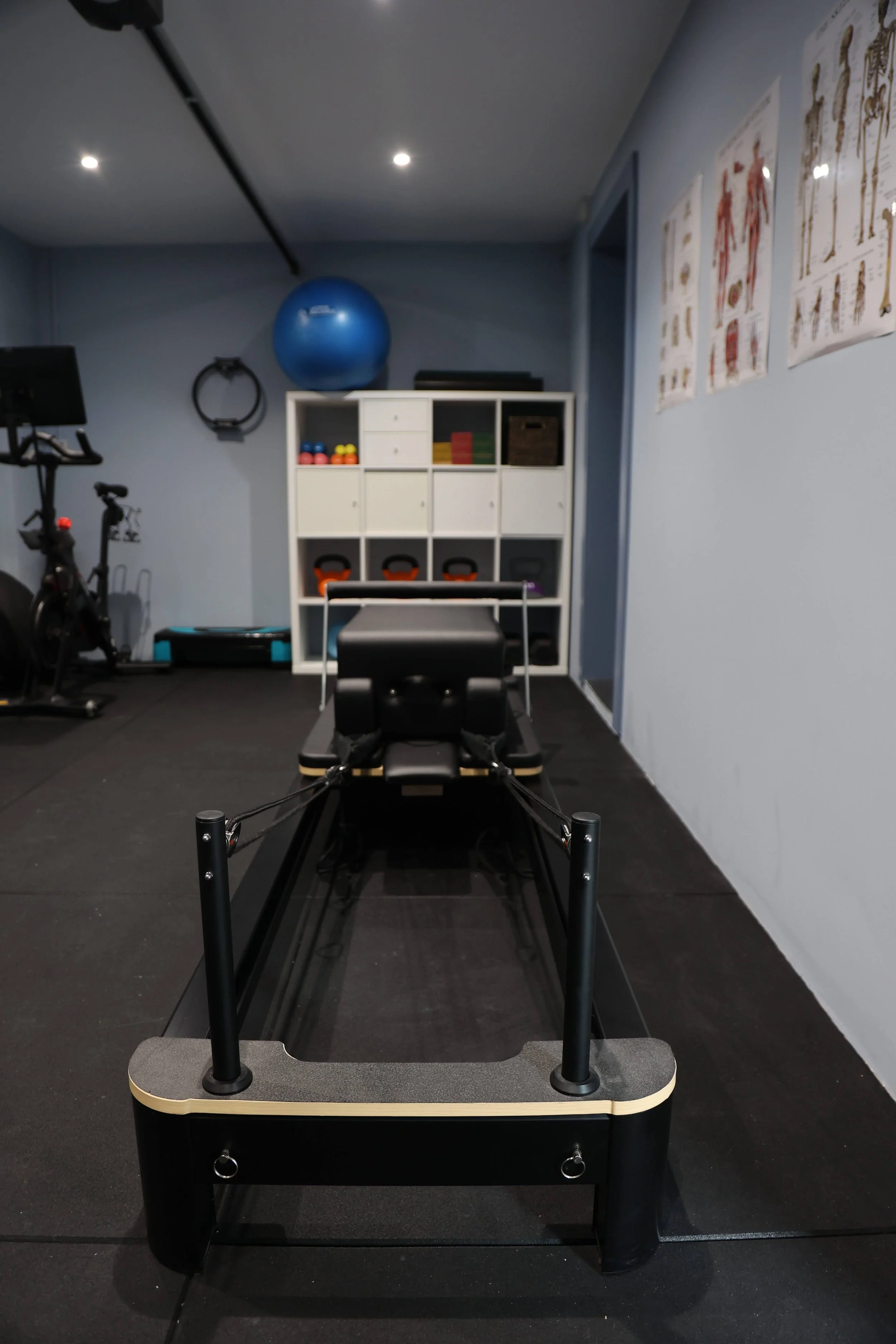 A reformer Pilates machine in a fitness room with workout posters on the wall, a large blue exercise ball, and various workout equipment.