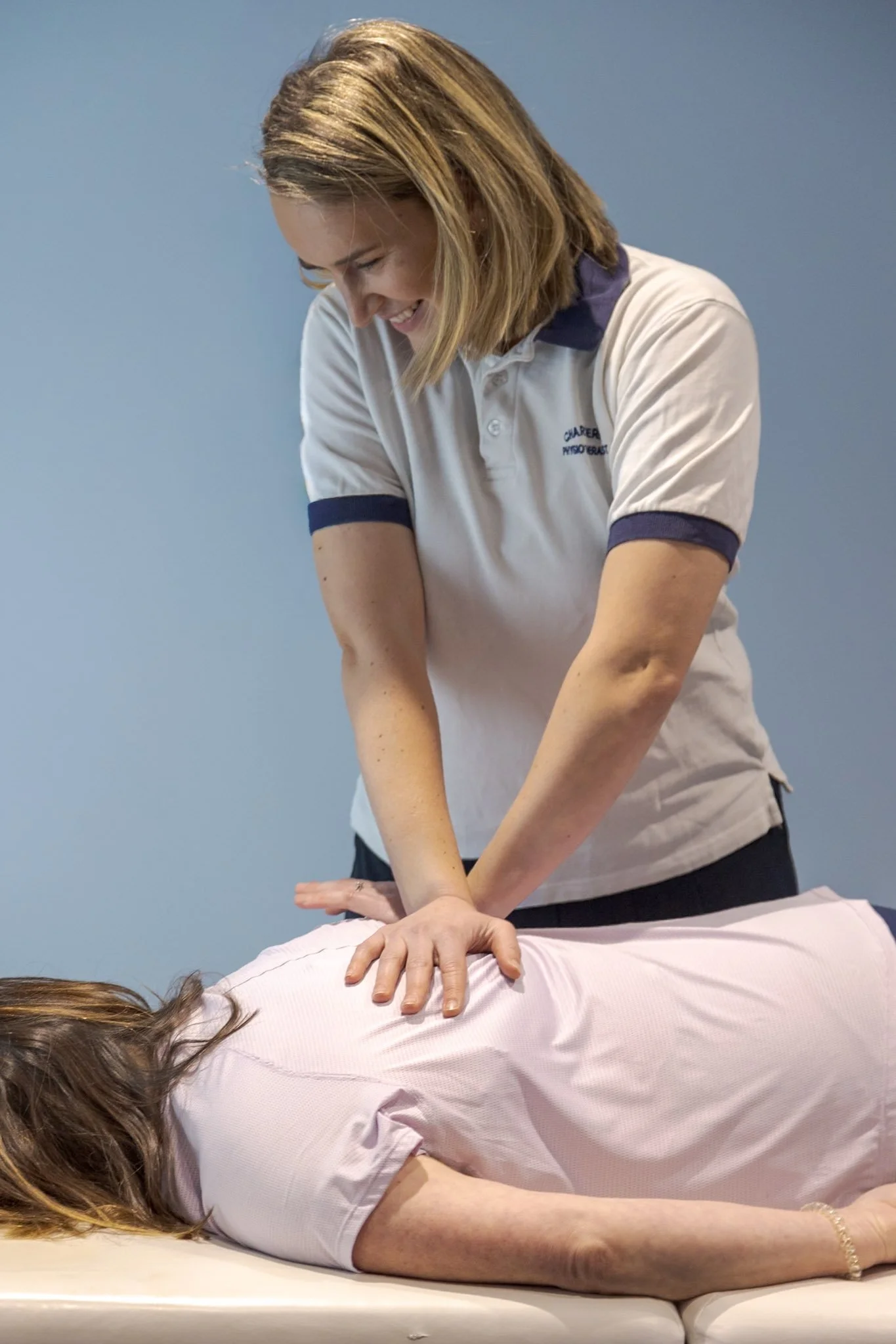 A woman wearing a white polo shirt giving a back massage to a person lying face down on a massage table, with a plain light blue background.
