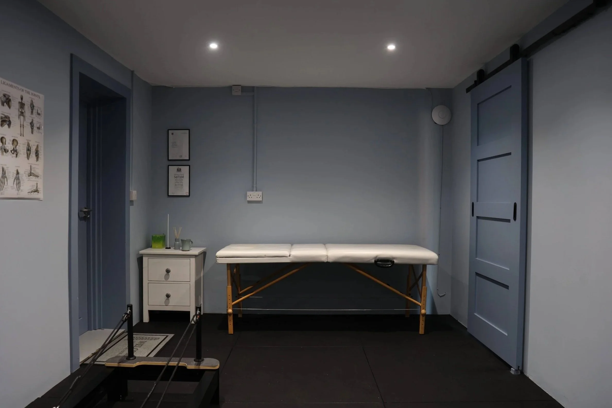 A small room with blue walls, a white bench, a white table with a small green box and some reed diffusers, framed certificates on the wall, a poster of the human skeleton, and equipment for a body assessment in the foreground.