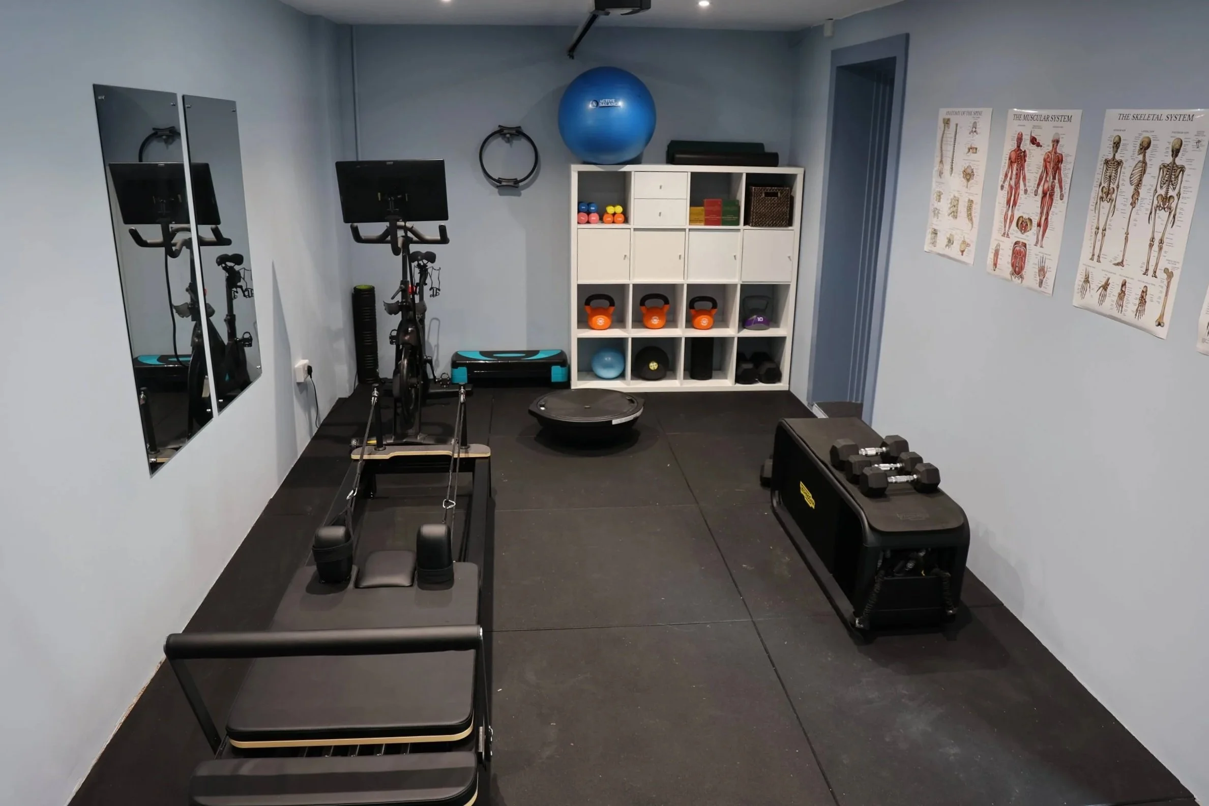 Home gym room with a stationary bike, kettlebells, a balance ball, medicine balls, resistance bands, and workout posters on the wall.
