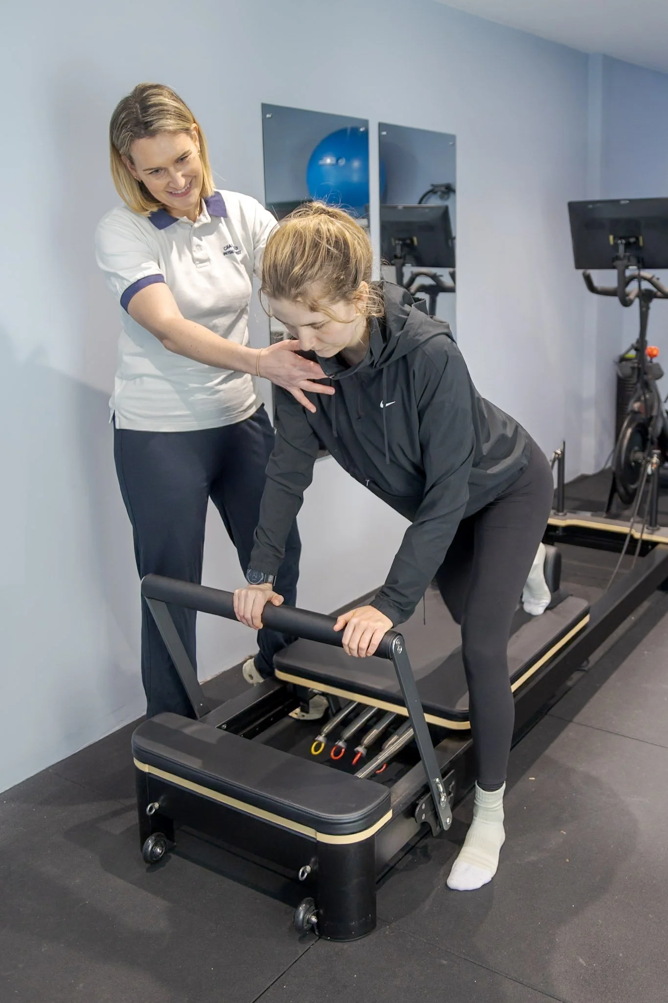 A woman and a girl are in a fitness or physical therapy setting with exercise equipment. The girl is performing a Pilates exercise on a reformer machine, holding the bar and leaning forward. The woman, standing beside her, is smiling and guiding her. The room has a light blue wall and some exercise bikes in the background.