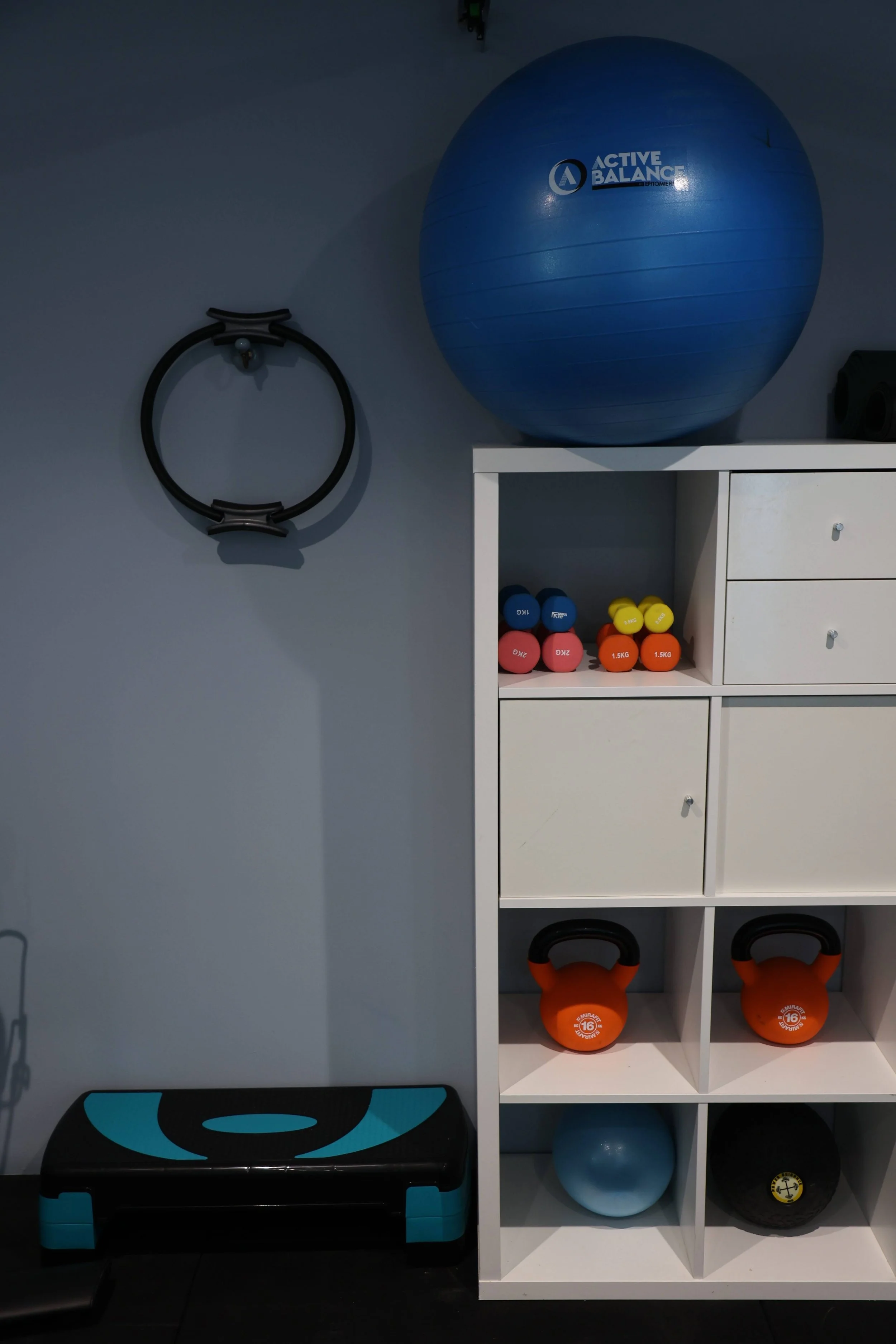 A blue stability ball on a white shelf, with a black kettlebell, colorful dumbbells, and black and orange kettlebells in the background, and a black and blue exercise step in the foreground.