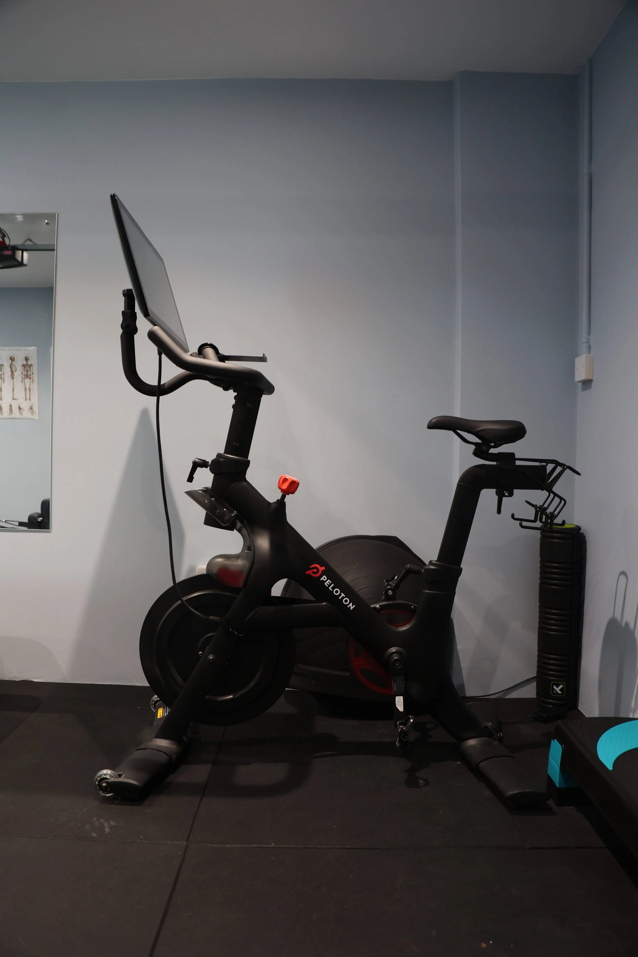 A black Peloton stationary exercise bike in a gym or workout room, with a digital screen on the handlebars, set against a light blue wall with a mirror and exercise poster, and a black foam roller on a rack nearby.