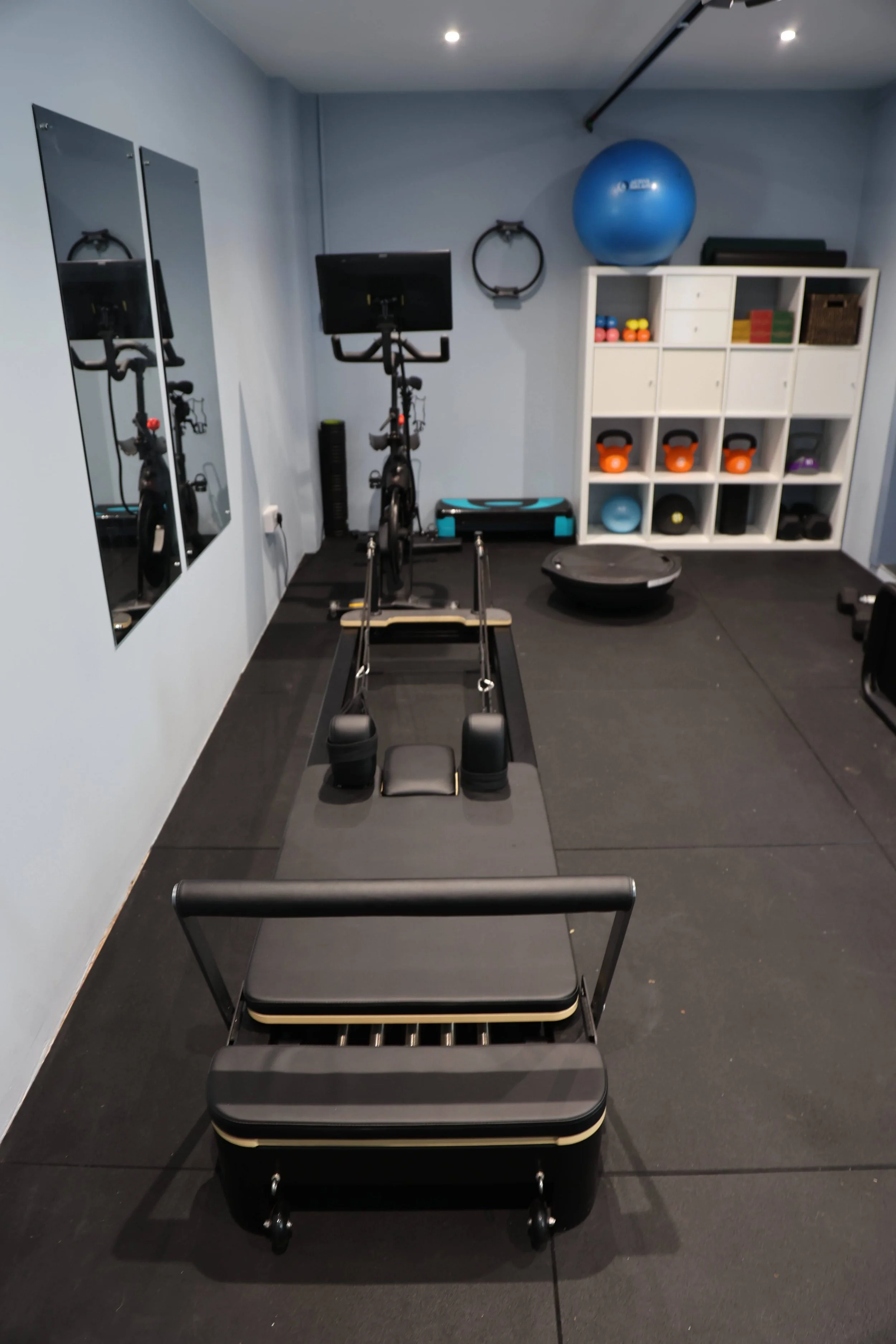 Home gym with a rowing machine on black rubber flooring, a full-length mirror on the left wall, and a white cubby shelf filled with exercise equipment like kettlebells, dumbbells, resistance bands, and a large exercise ball, under a sloped ceiling wi