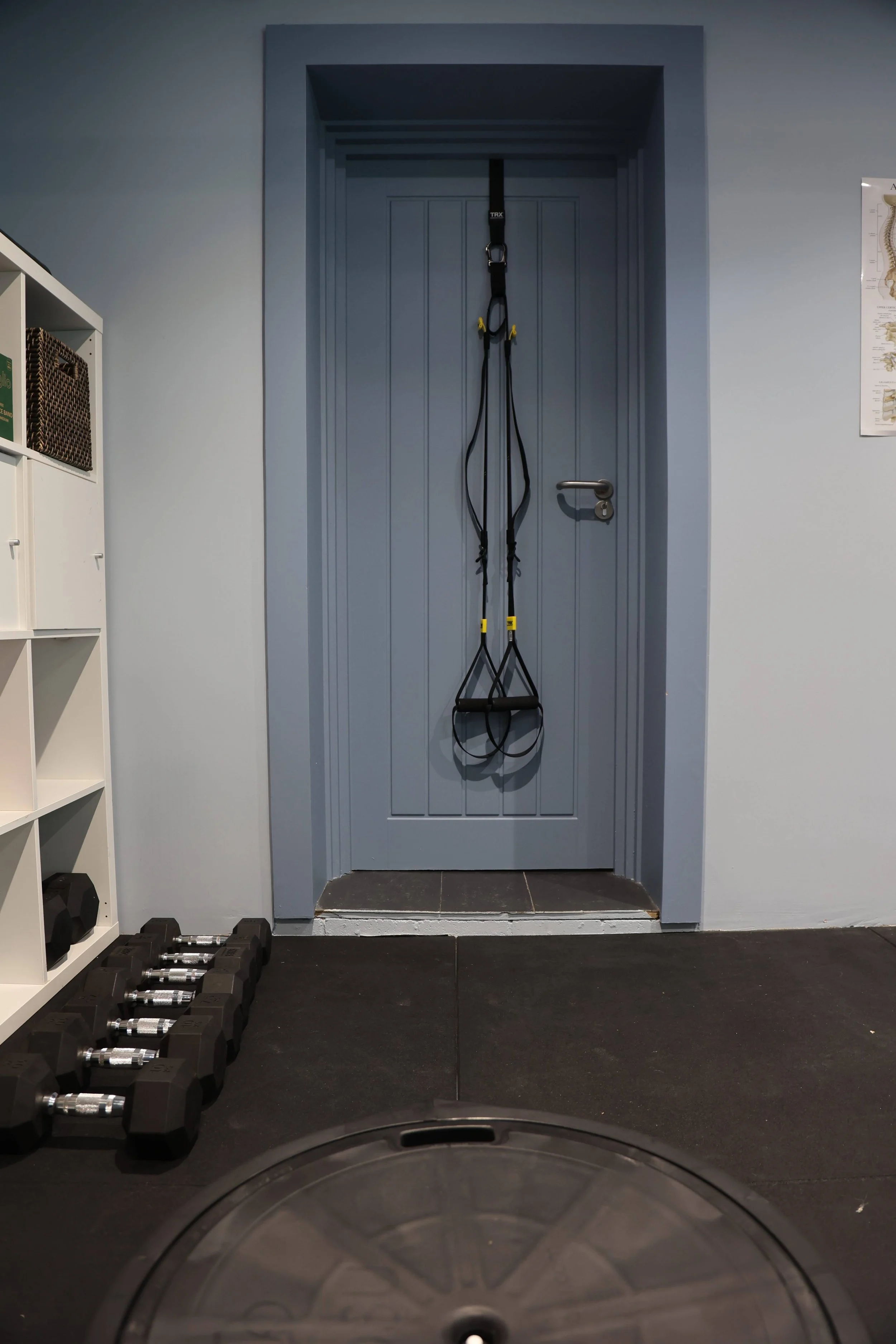 View inside a gym, showing a blue door with a suspension trainer hanging on it, dumbbells on the black rubber floor, and a white storage shelf with baskets on left side.