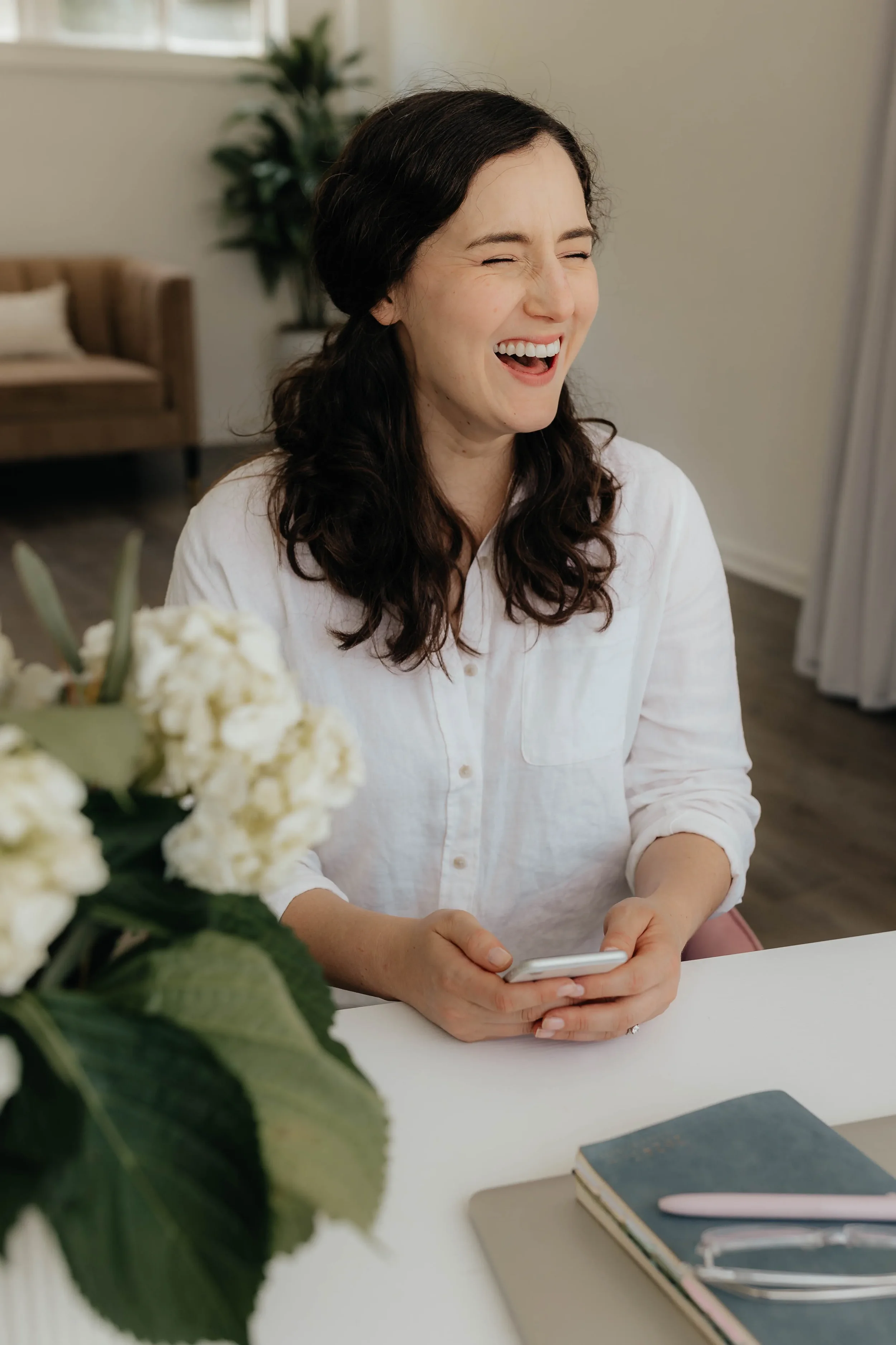 A woman with dark wavy hair, wearing a white shirt, is laughs and holds her phone, sitting at a white table with a bouquet of white flowers and notebooks in front of her.