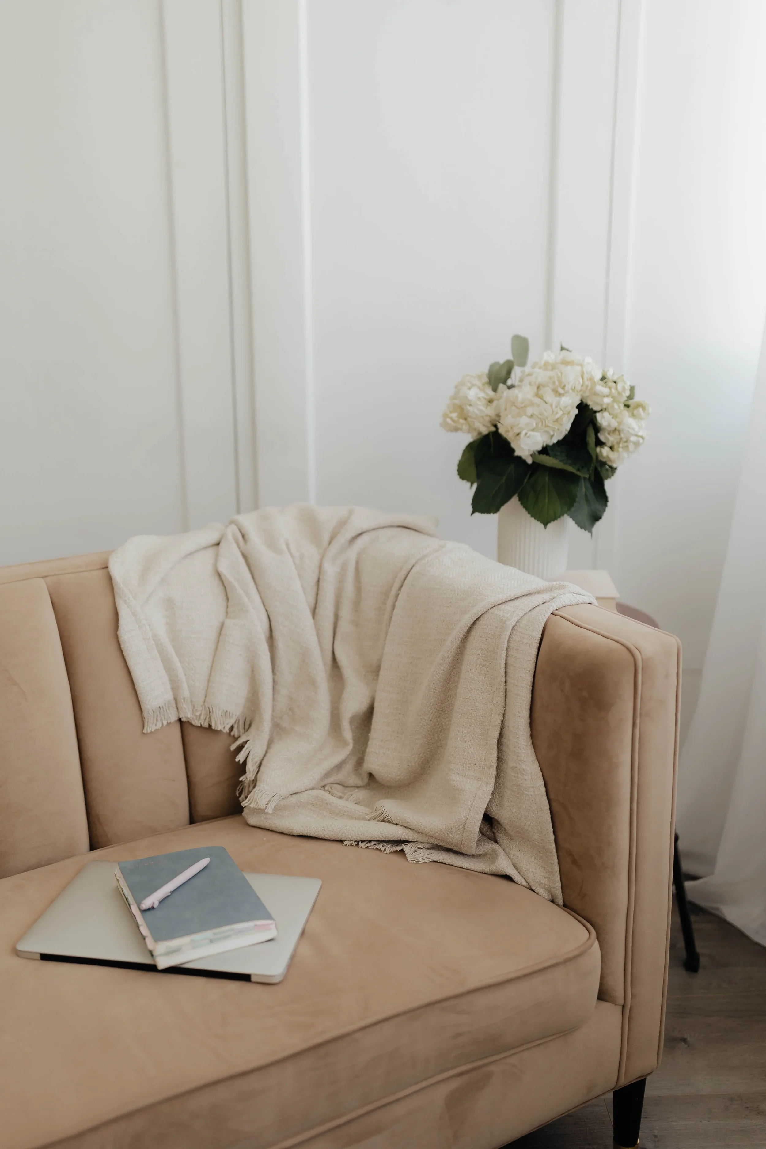 A beige sofa with a cream-colored blanket draped over the armrest. On the sofa's wooden armrest, there's a closed laptop with a notebook and a pen on top. In the background, a white vase with white flowers and dark green leaves is placed on a white side table against white paneled walls.