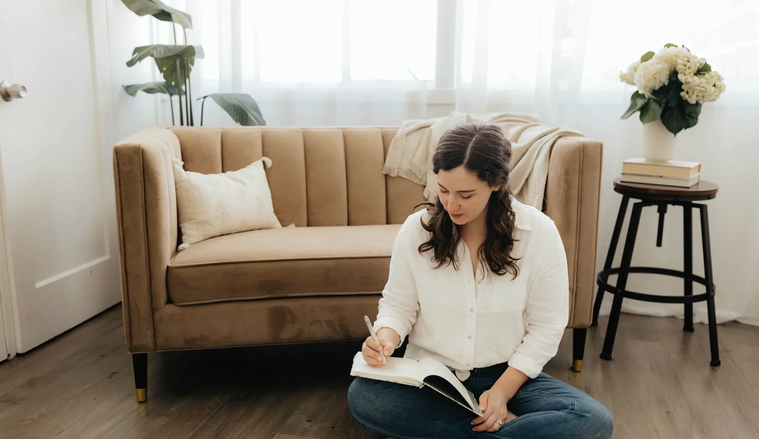 A woman with long dark hair, dressed in a white shirt and jeans, sitting on the floor and writing in a notebook.