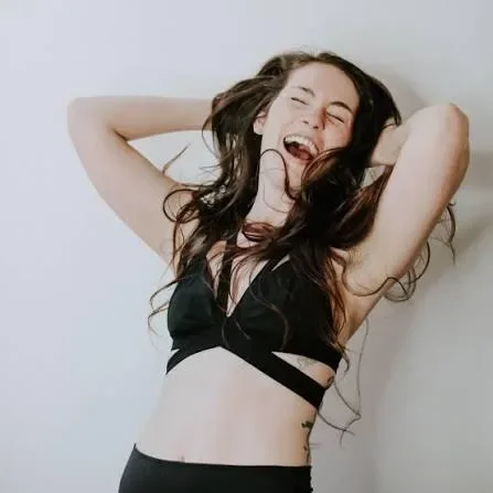 A young woman with long dark hair laughing and posing with her arms behind her head against a plain white wall.