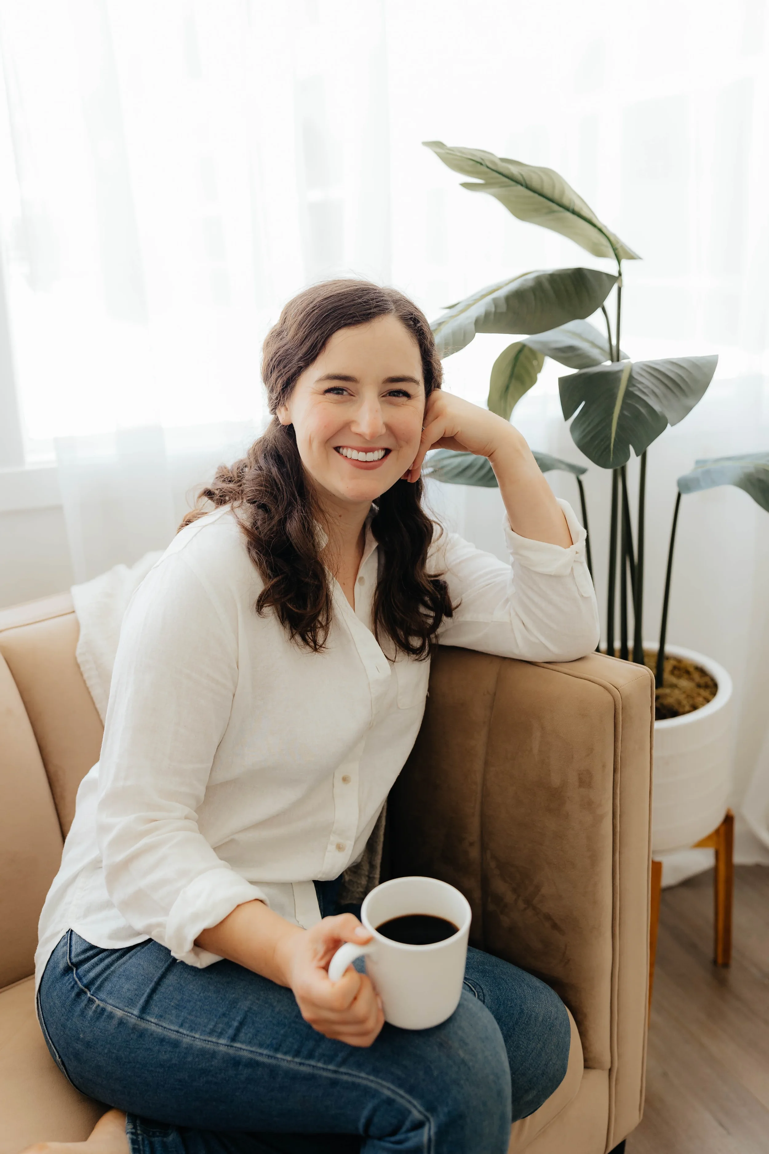 A woman sitting on a beige couch holding a mug of coffee, smiling, with a large green plant and white curtains in the background.