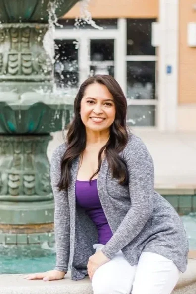 A smiling woman with long dark hair, wearing a purple top and gray cardigan, sitting by a fountain outdoors.