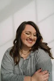 A woman with long hair, glasses, and a gray sweater smiling and looking to the side indoors.