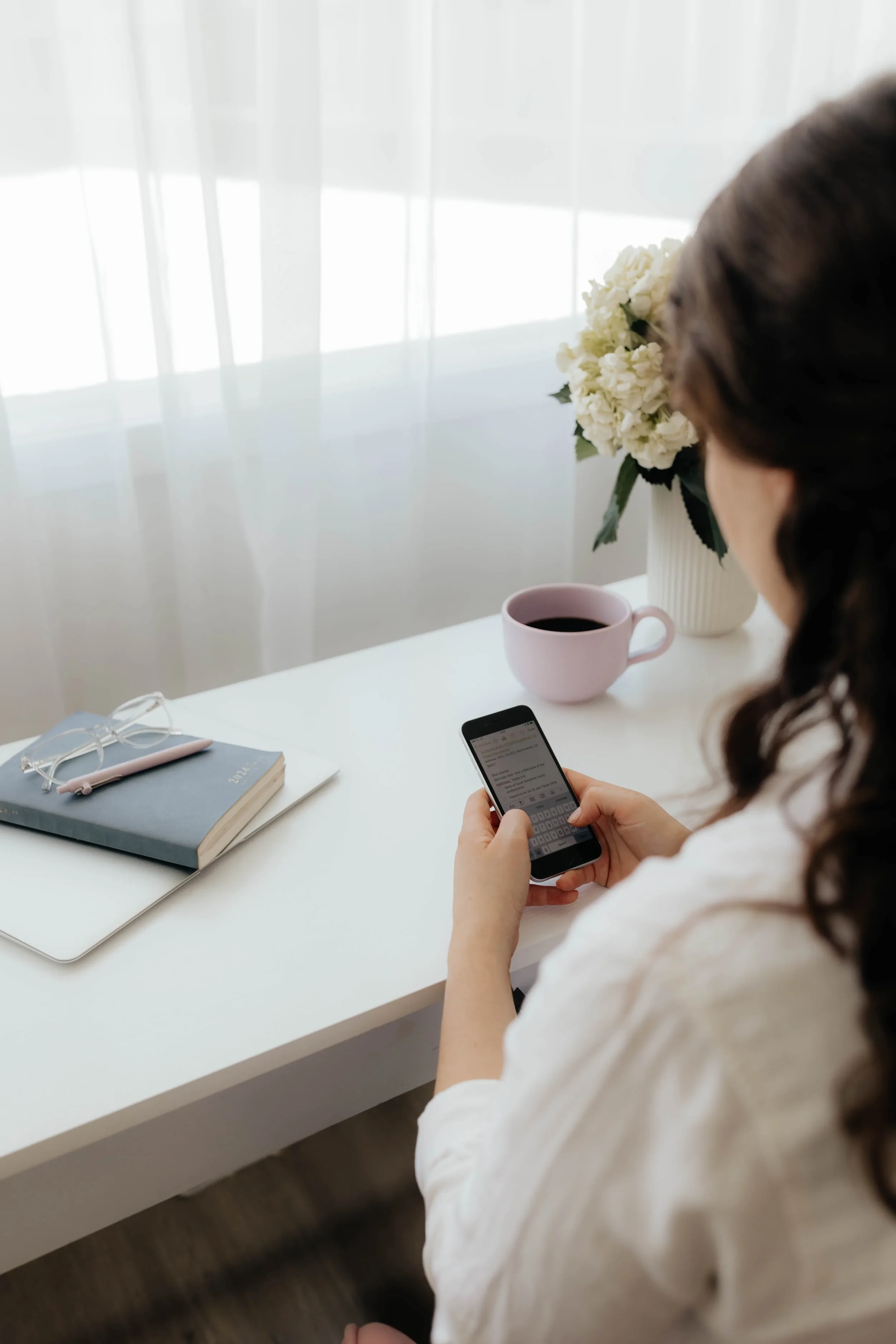 A woman sitting at a white desk looking at her smartphone, with a pink mug, a closed notebook with glasses and a pen on top, and a vase with white flowers in the background, near sheer white curtains.