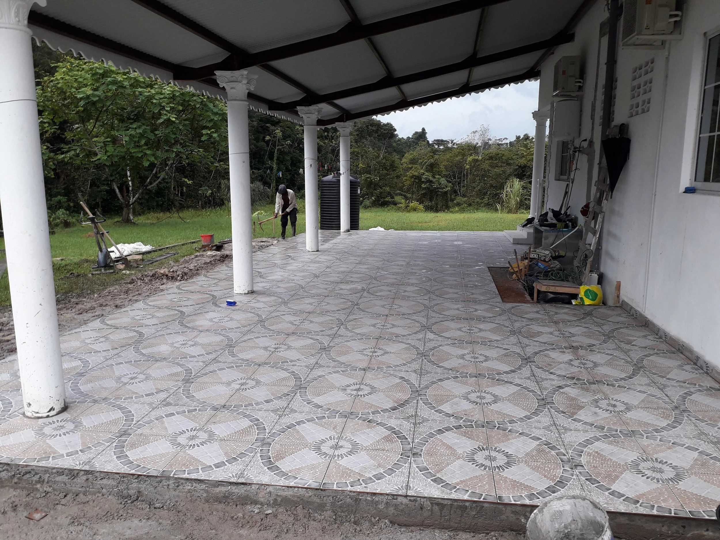 Une terrasse couverte avec des piliers blancs, en train de recevoir des carreaux de sol avec un motif géométrique. Un homme travaille à l'extérieur dans une zone herbeuse, avec des arbres en arrière-plan. Des outils et du matériel de construction sont visibles contre le mur de la maison.