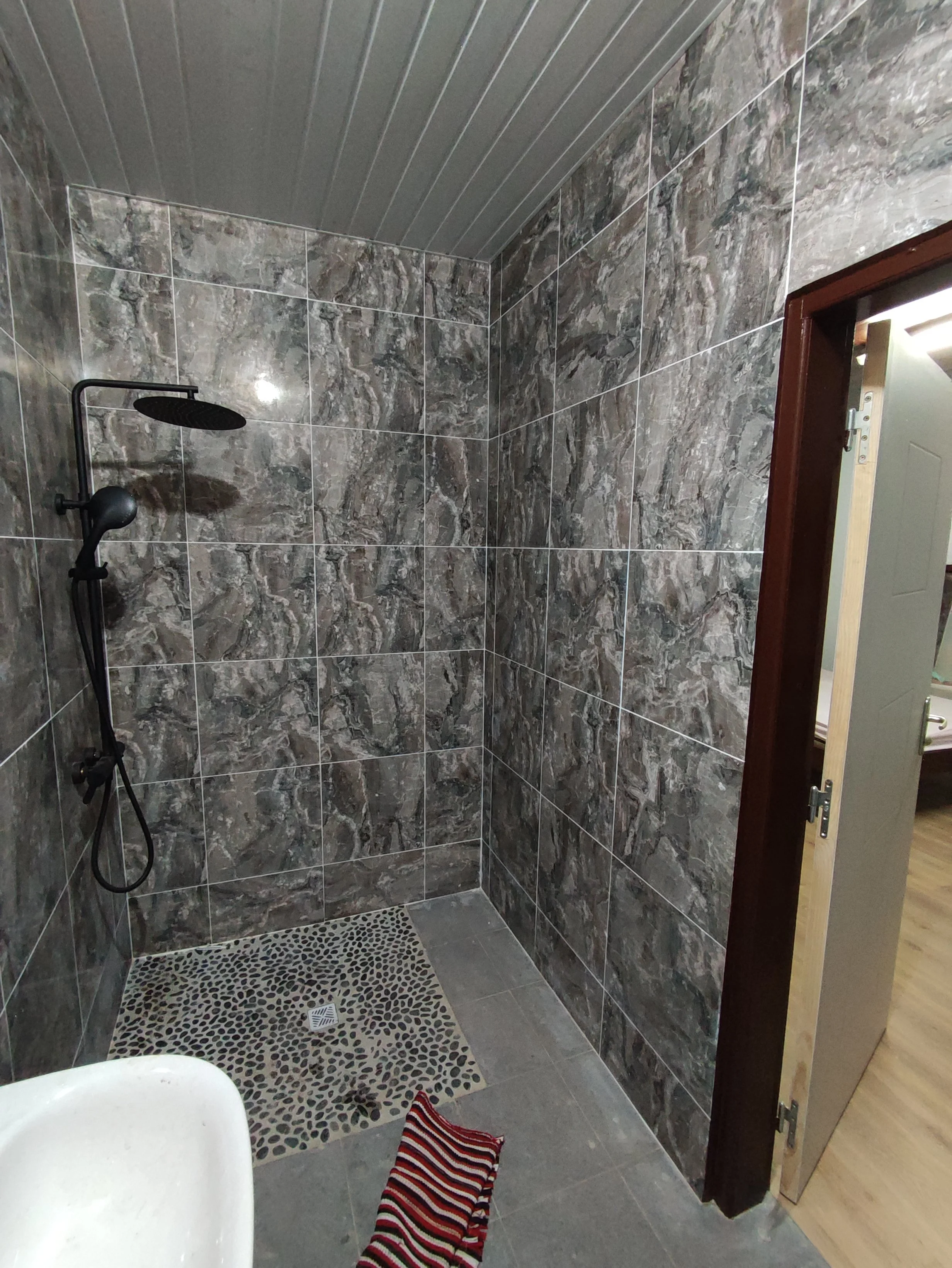 Salle de bain avec une douche à l'italienne, murs en marbre, sol gris, un tapis à rayures, se voit également un lavabo partiel en bas, et un mur en bois autour de la porte.