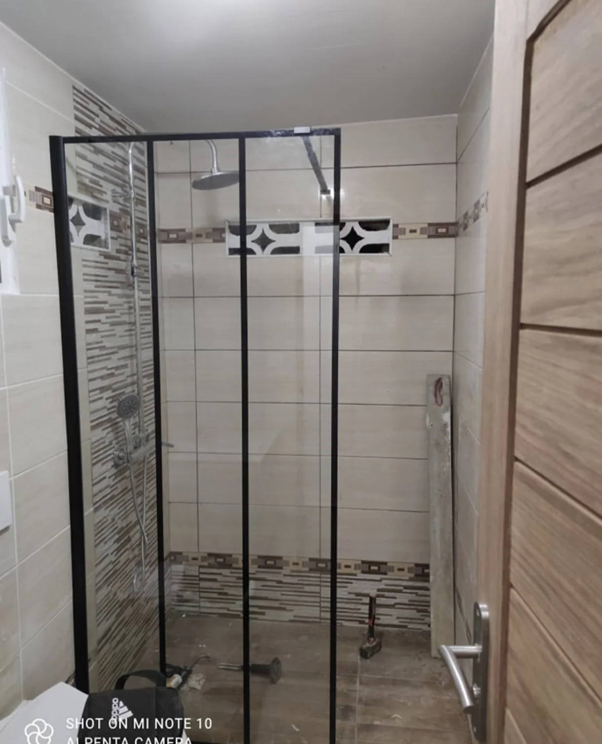 Une cabine de douche en construction avec un receveur, un receveur de douche, des murs en carrelage beige avec motifs et un porte en verre avec cadre noir.