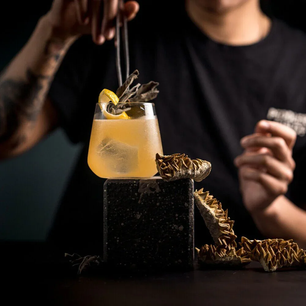 A person holding a cocktail glass with a yellow drink, lemon slice, and dried garnish, surrounded by dried artichokes, on a black surface with a dark background.