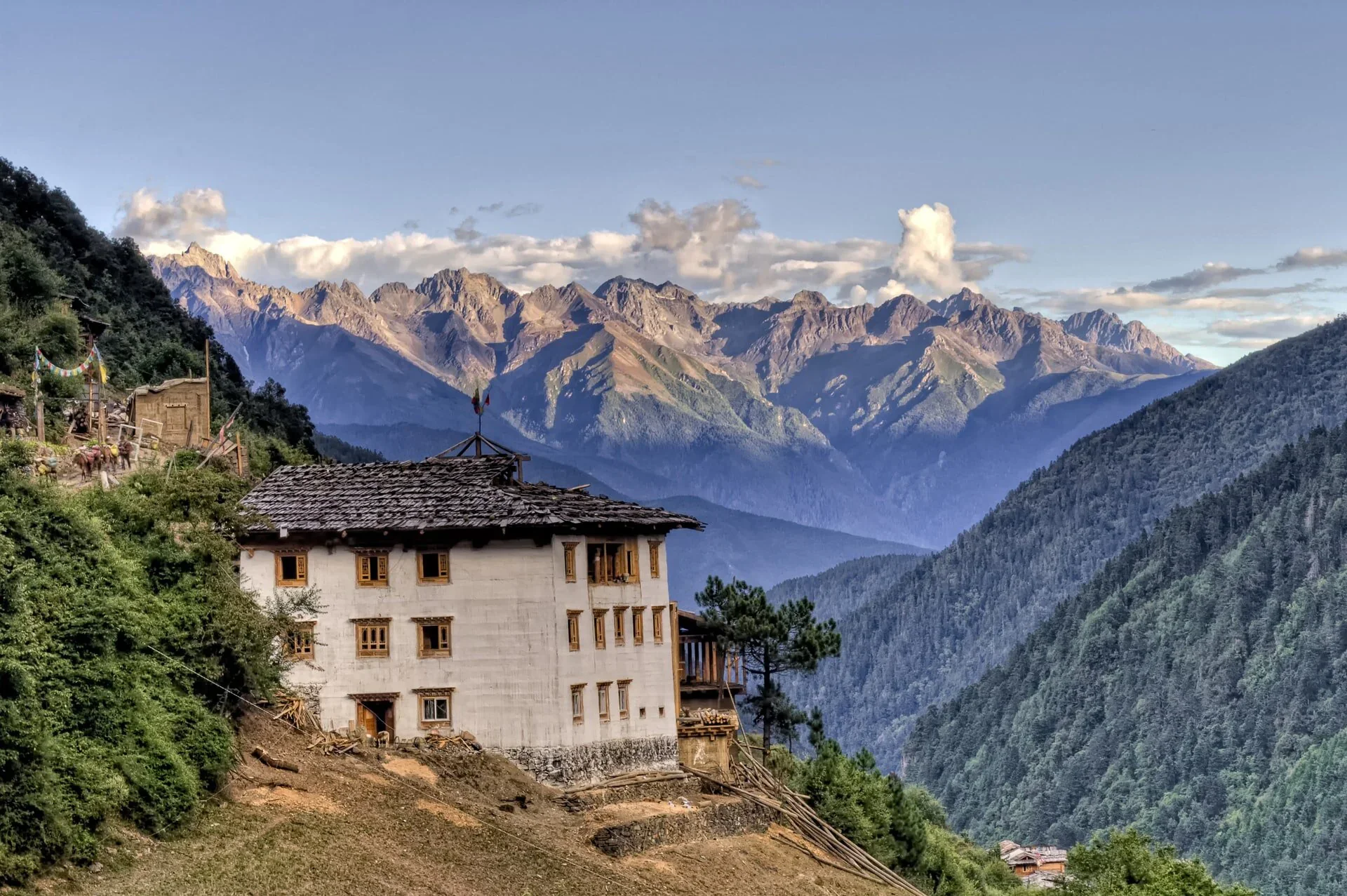 Mountain village in Sichuan, China with a white building with wooden windows, surrounded by green forested hills and tall mountain peaks in the background under a partly cloudy sky.