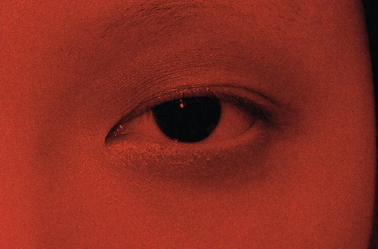 Close-up of a human eye with a dark pupil and eyelid, ambient lighting creating a reddish hue.