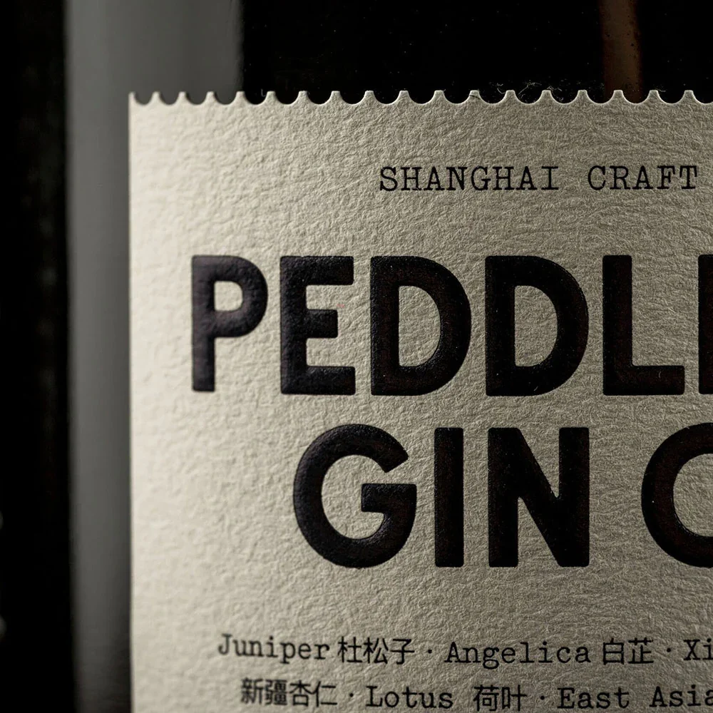 Close-up of a textured label on a Peddlers Shanghai craft gin bottle, highlighting the words "Peddlers Gin" and ingredients listed in English and Chinese.