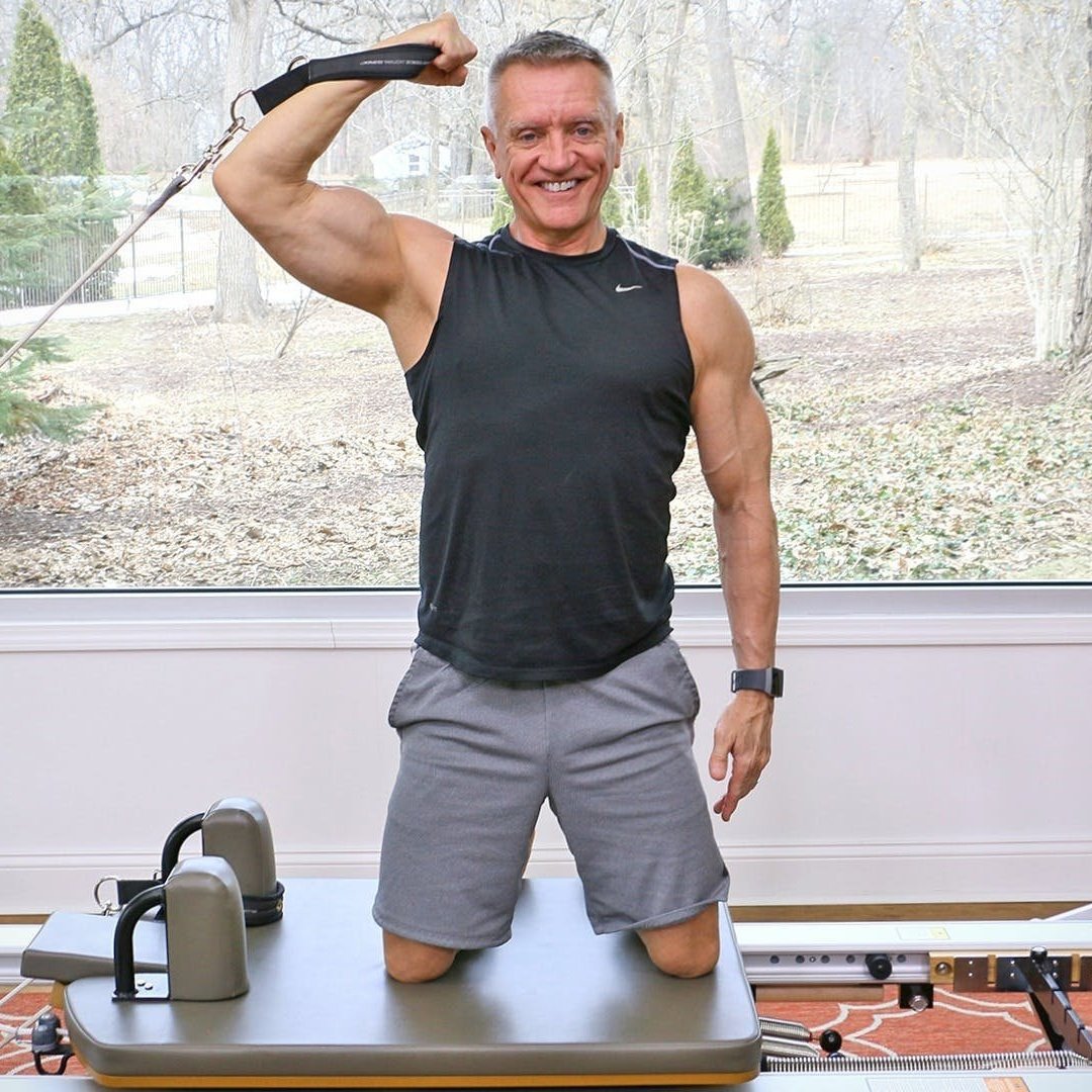 Man kneeling on a treadmill in a room with large window, wearing a black sleeveless athletic shirt and gray shorts, smiling and holding a resistance band over his shoulder, with kettlebells nearby.