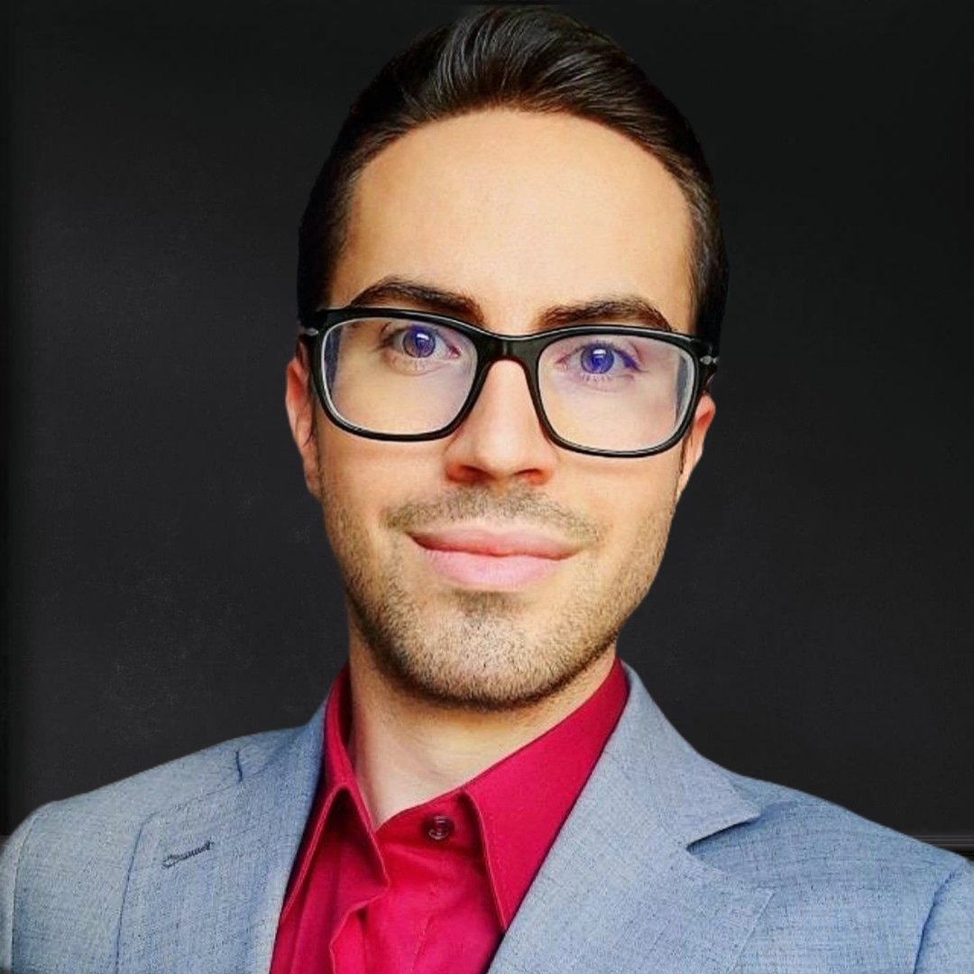 Porträt eines Mannes mit dunklem Haar, Brille, grauem Anzug und rotem Hemd vor dunklem Hintergrund.