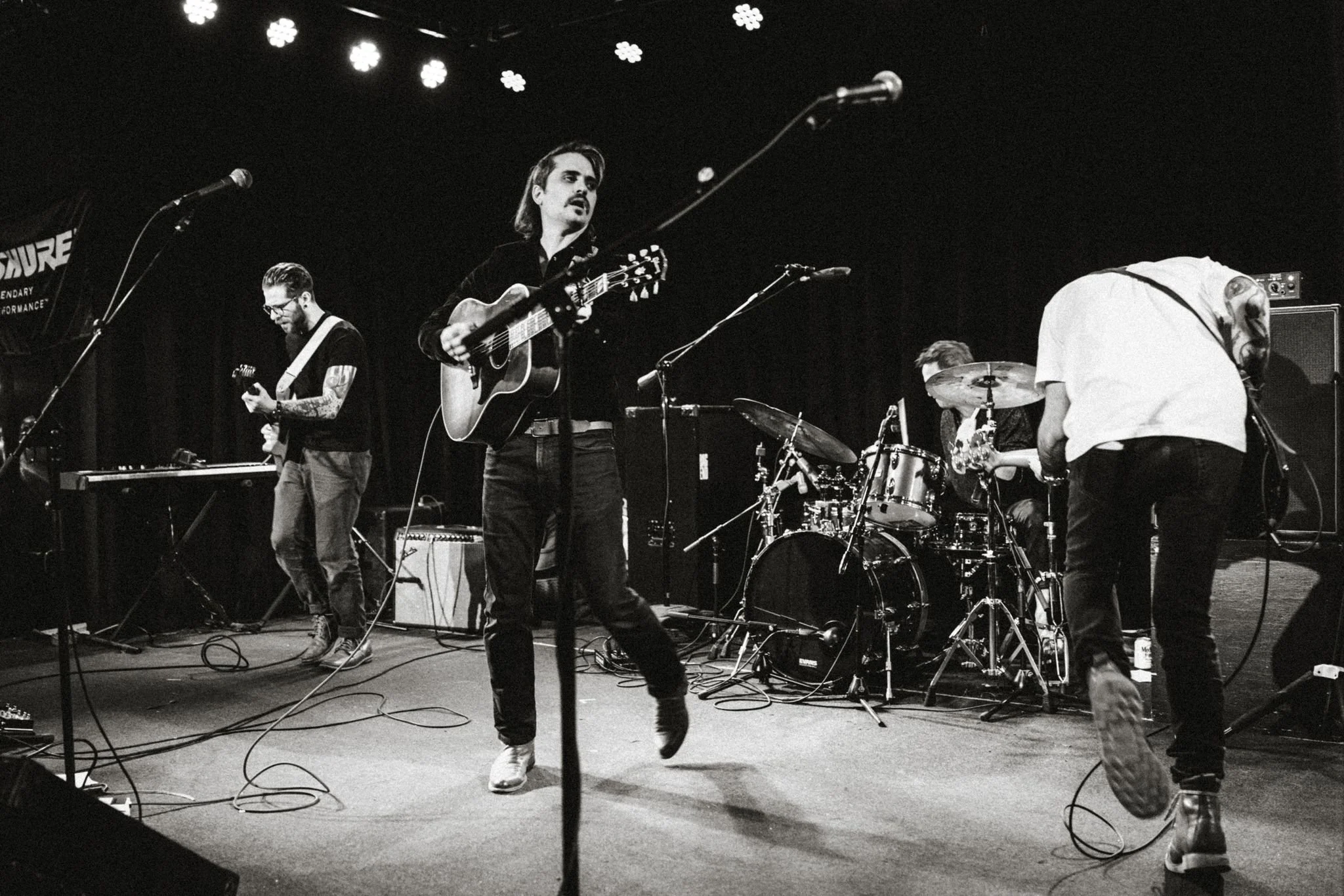 A black-and-white photo of a four-piece band performing on stage, with a drummer, guitarist, and two other musicians, one of whom is wearing glasses and tattoos, under stage lights.