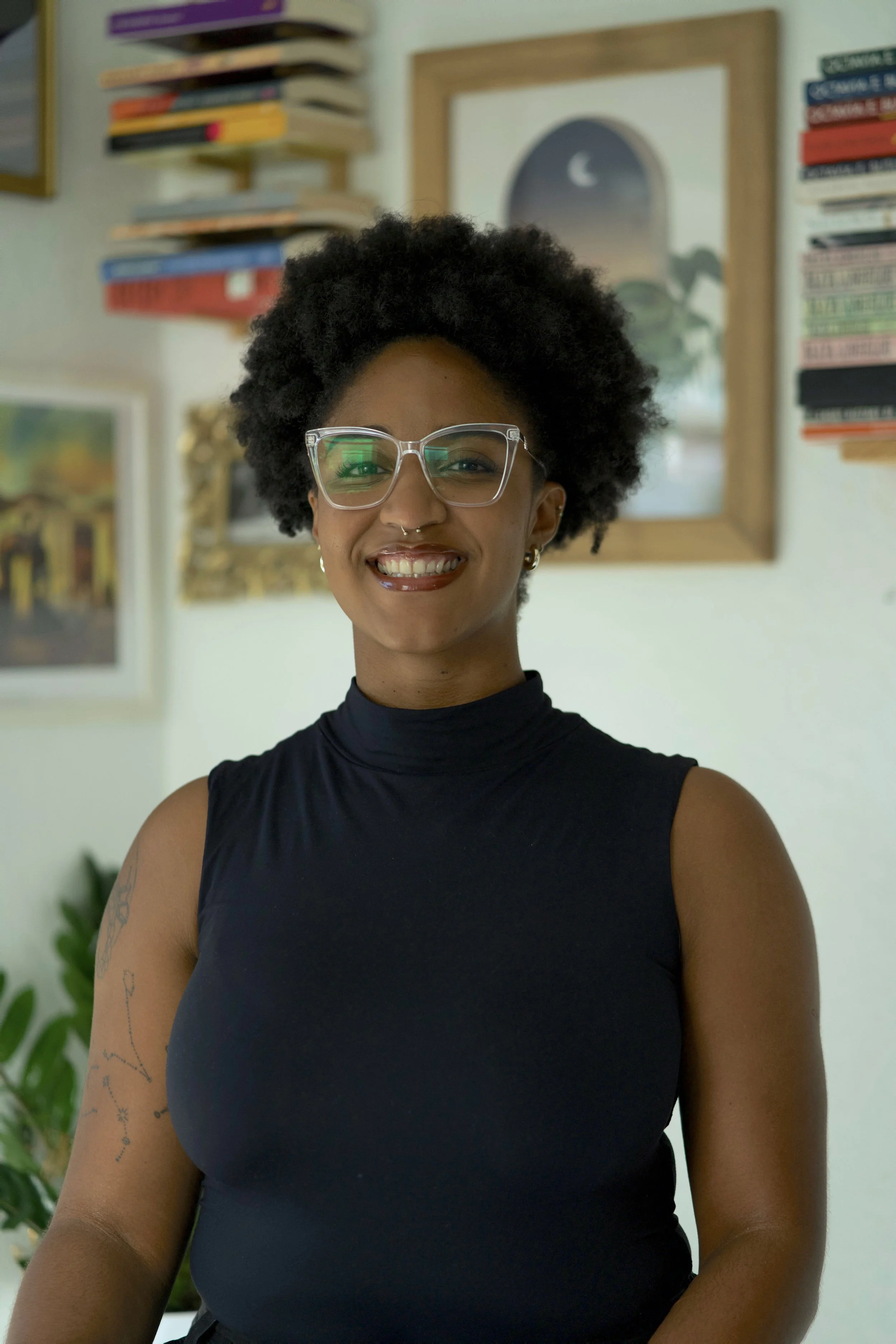 Portrait of Makayla with glasses and a tattoo on her left arm, smiling in a room with books and artwork on the wall.