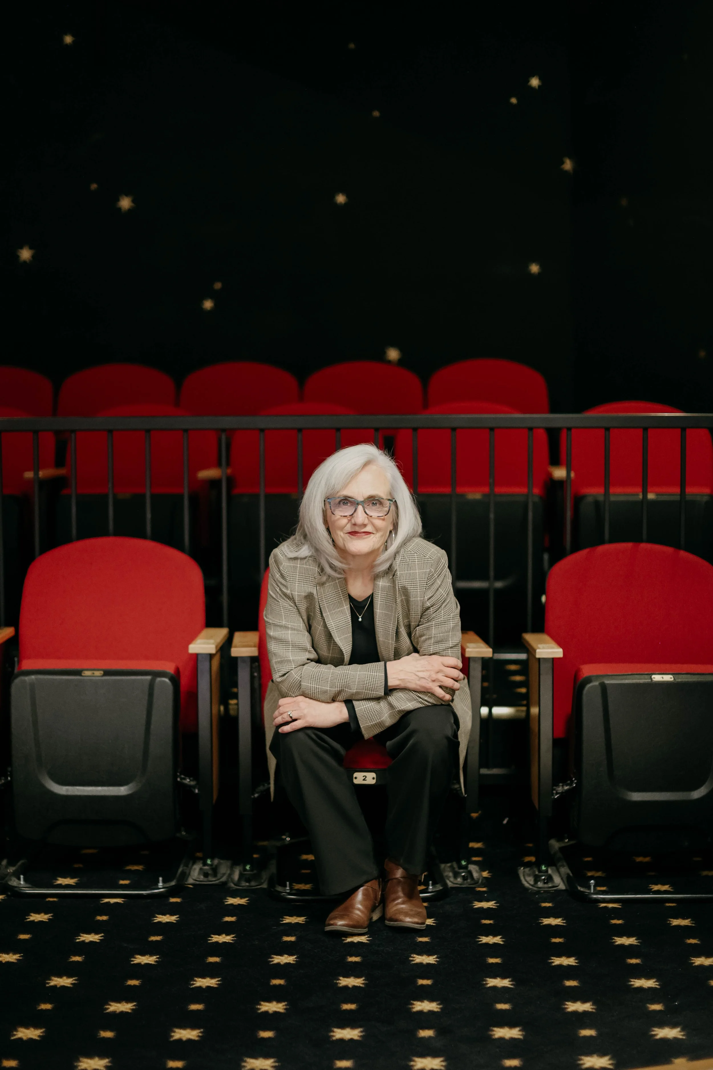 An elderly woman with white hair, glasses, and a beige checkered blazer sitting alone in a movie theater with red seats and a starry night sky mural on the wall behind her.