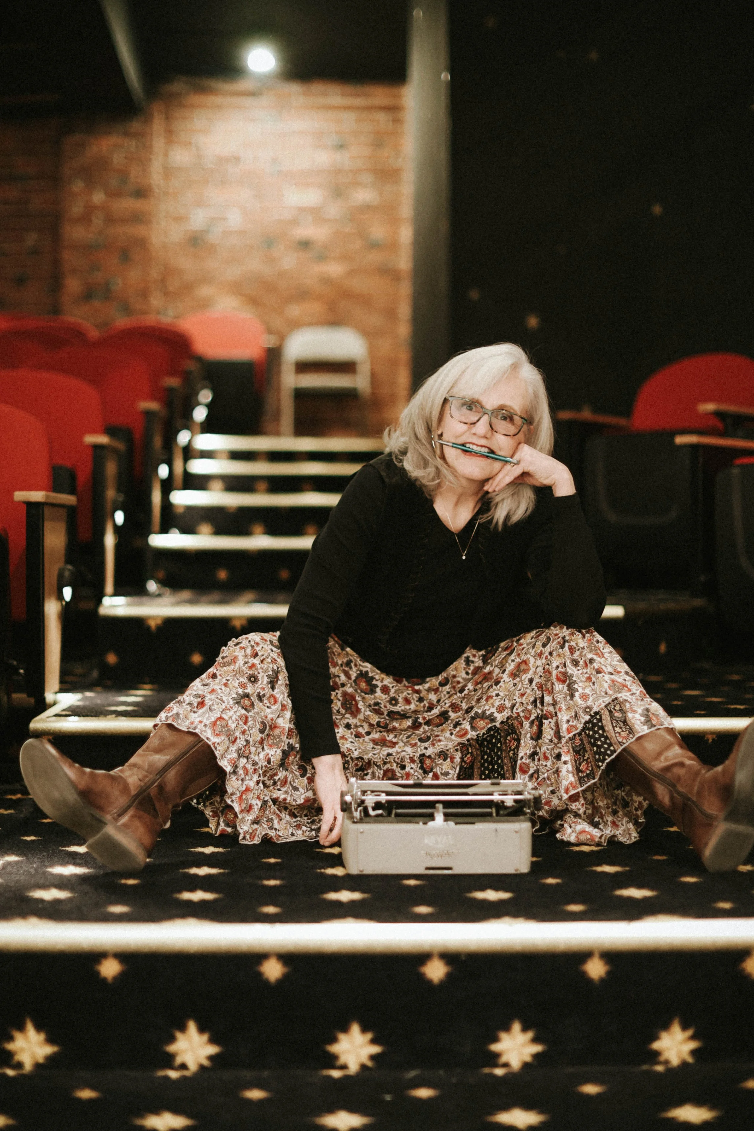 A woman sitting on a theater aisle floor, smiling, with a vintage typewriter in front of her, holding a pen in her mouth, in a theater with red seats and a brick wall background.