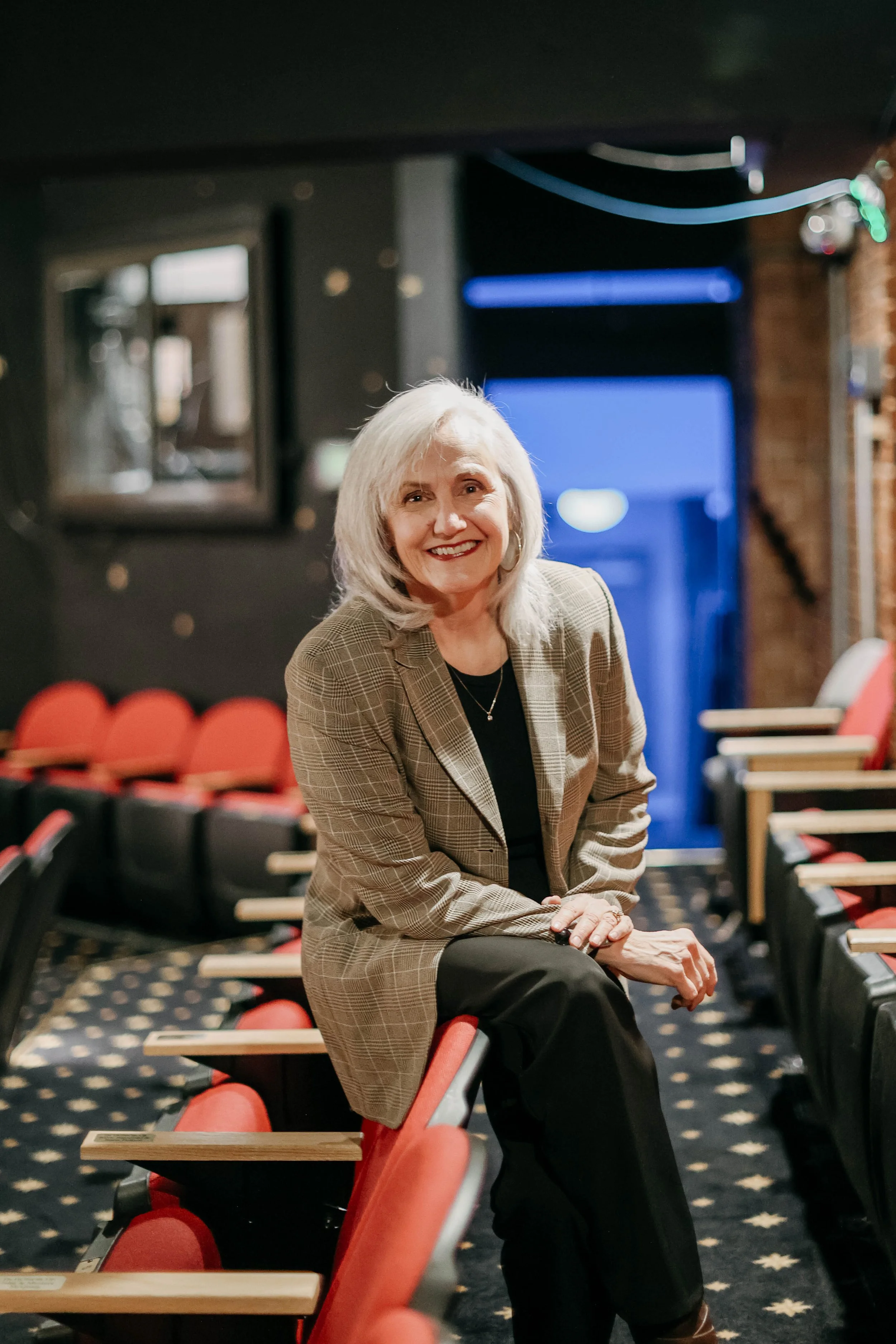 A woman with gray hair smiling, sitting on a theater seat in an auditorium with red seats and a patterned carpet.
