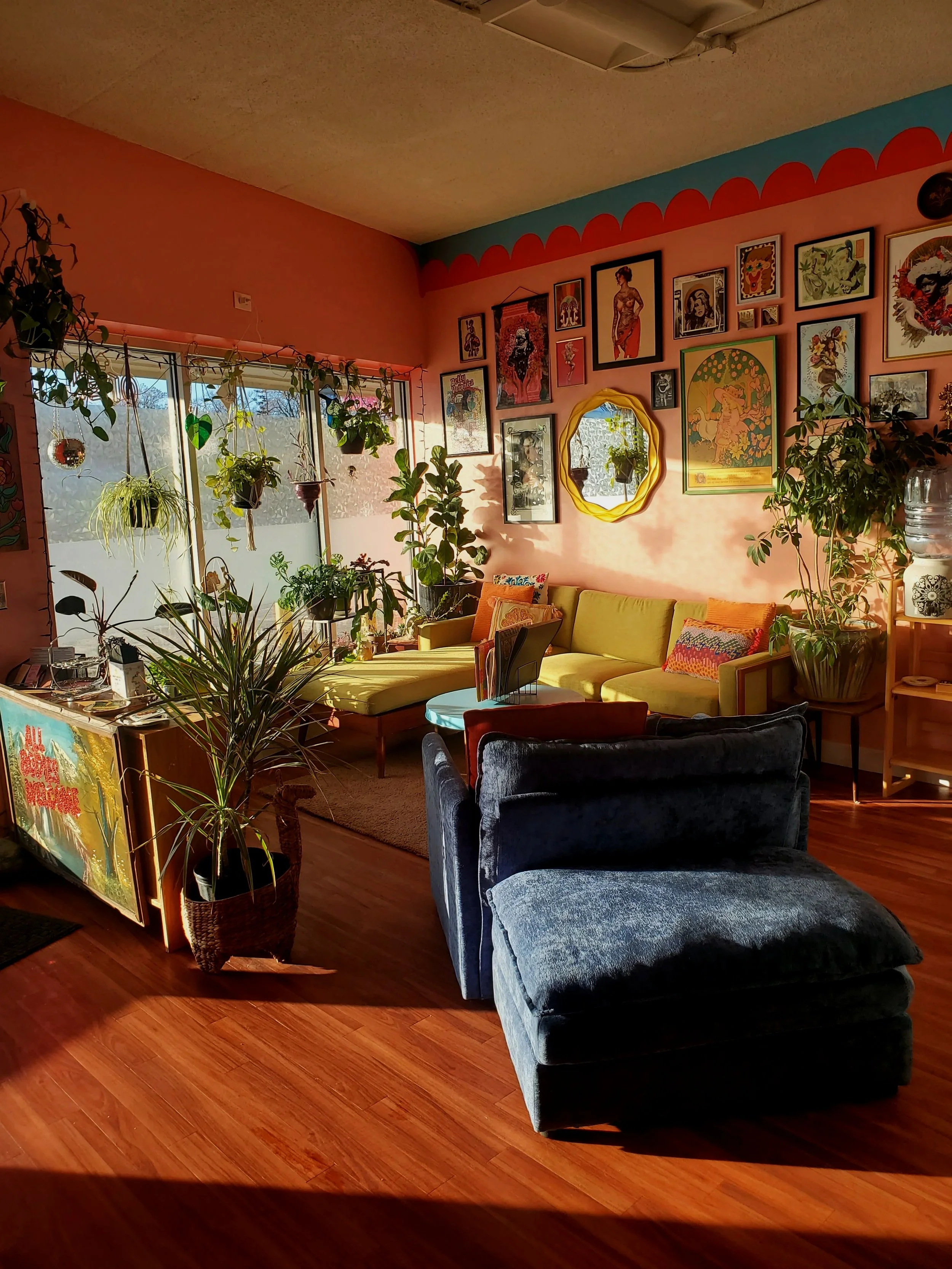 Colorful tattoo shop waiting area with a green couch, framed tattoo art gallery wall, indoor plants, and a welcoming front counter displaying stickers and merchandise.